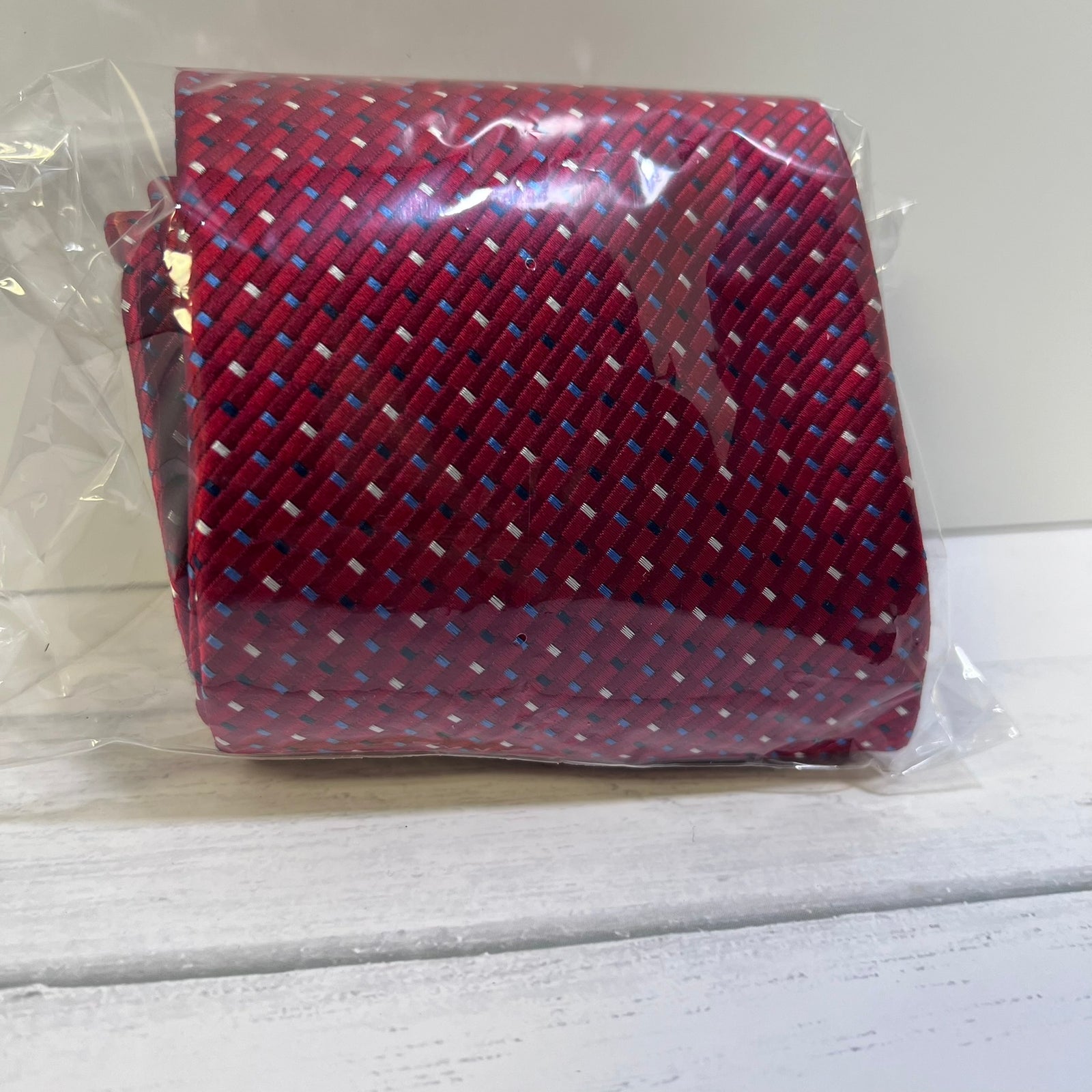 A rolled necktie in clear plastic packaging, featuring a red base with a woven pattern of blue, white, and black threads.