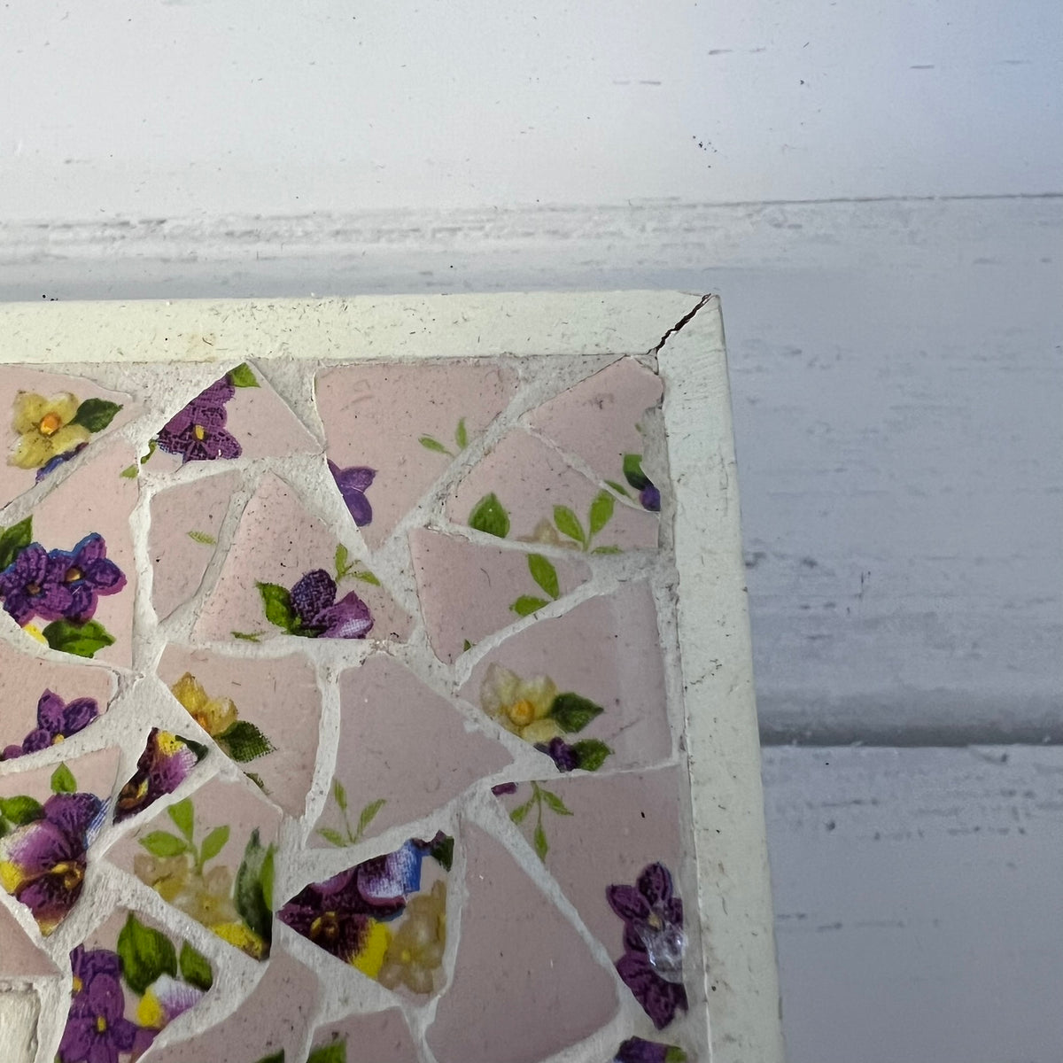 A close-up of a square mosaic tile piece with a white distressed border, featuring pink tiles and a floral pattern of purple pansies, yellow flowers, and green leaves, set against a white painted wood background.