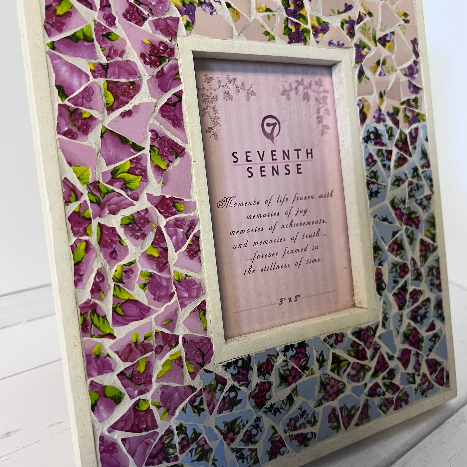 A square framed mosaic photo frame featuring purple, pink, and blue glass tile patterns around a central light pink insert with the text 'SEVENTH SENSE' and a small logo, shown on a white wood background.