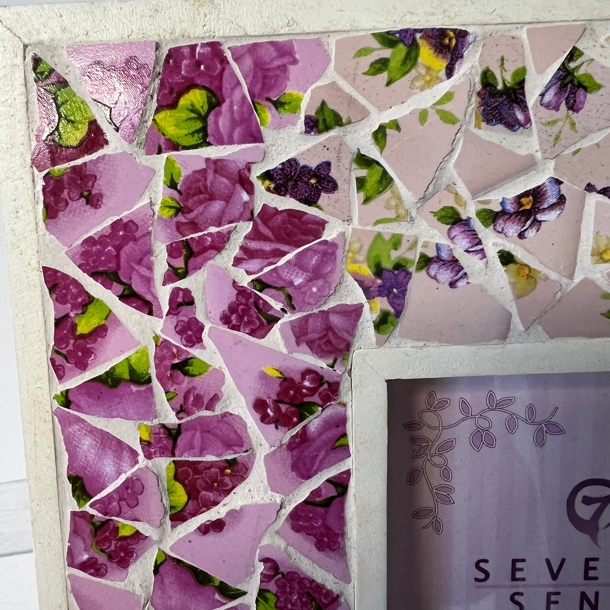A close-up of a purple floral mosaic tile wall with broken ceramic pieces in shades of pink, purple, and green, mounted on a light-colored frame or board. Part of a sign reading 'SEVEREN' is visible in the lower right corner.