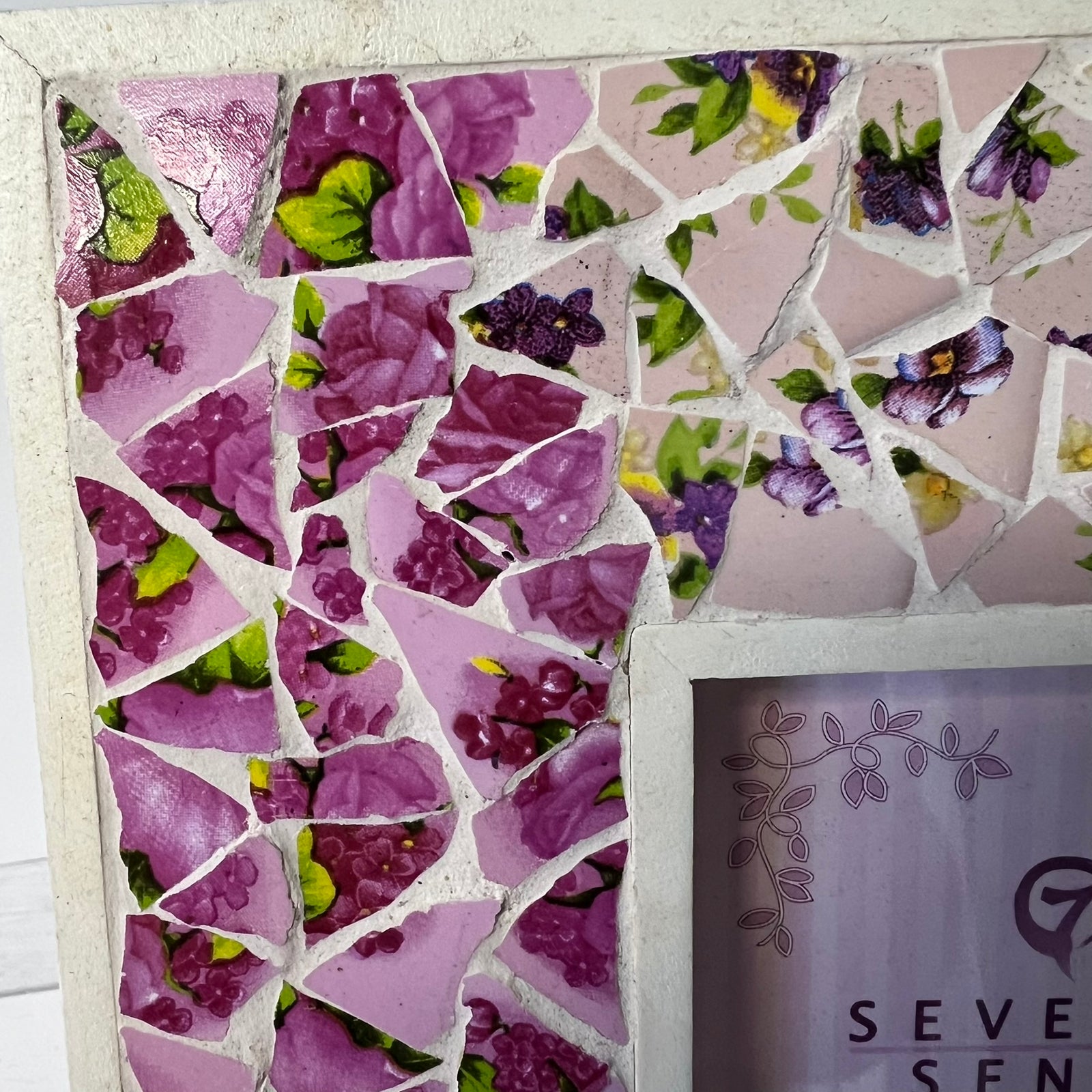 A close-up of a purple floral mosaic tile wall with broken ceramic pieces in shades of pink, purple, and green, mounted on a light-colored frame or board. Part of a sign reading 'SEVEREN' is visible in the lower right corner.