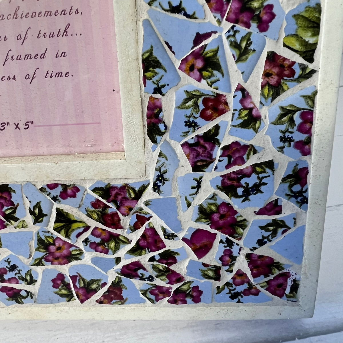 A close-up of a framed mosaic tile artwork featuring light blue background tiles with pink and purple floral patterns, white grout lines, and a light pink framed corner with cursive text.
