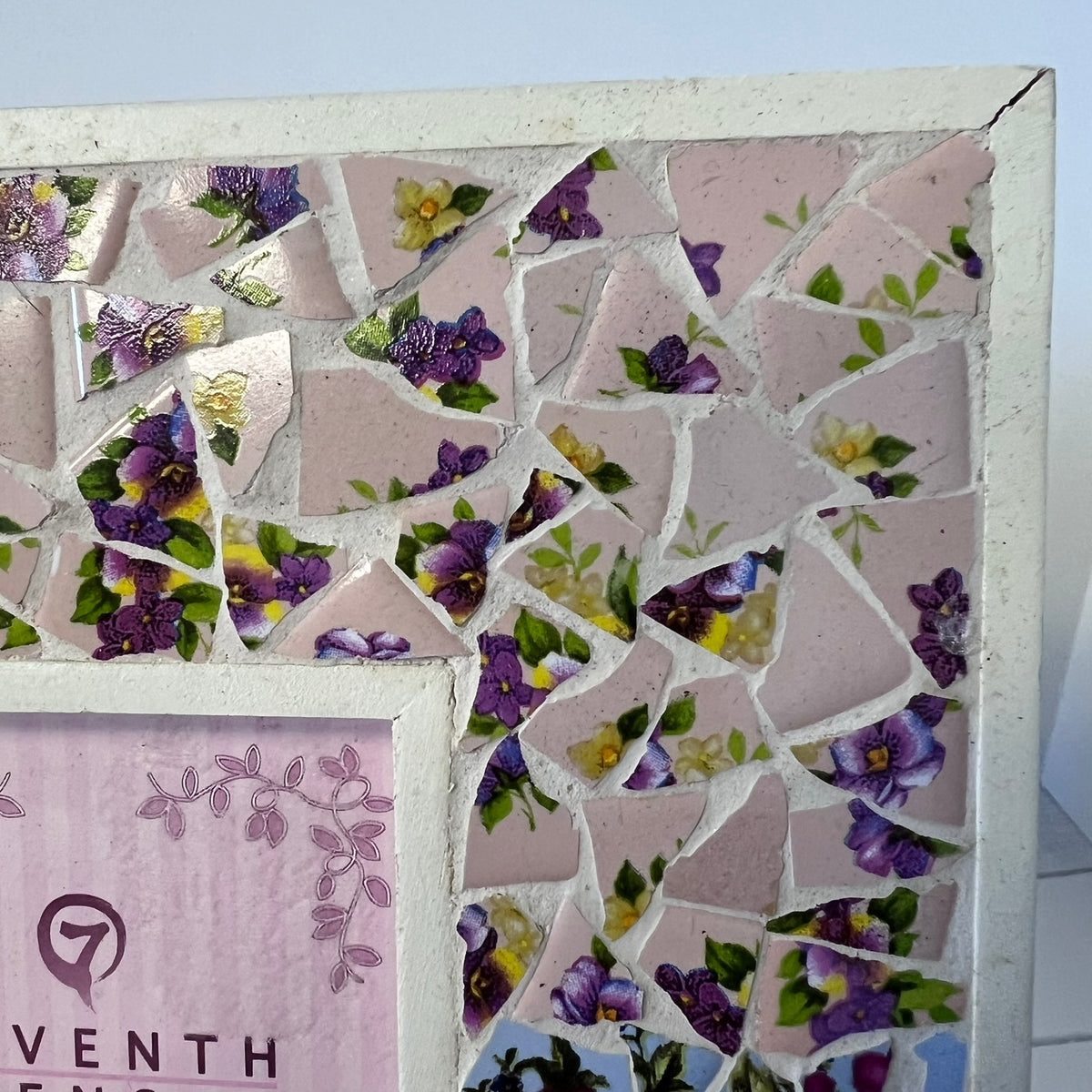 A close-up of a mosaic tile wall art piece featuring pink square tiles with printed purple and yellow flowers, green leaves, and white grout lines, with a white frame border and part of a pink decorative inset visible.