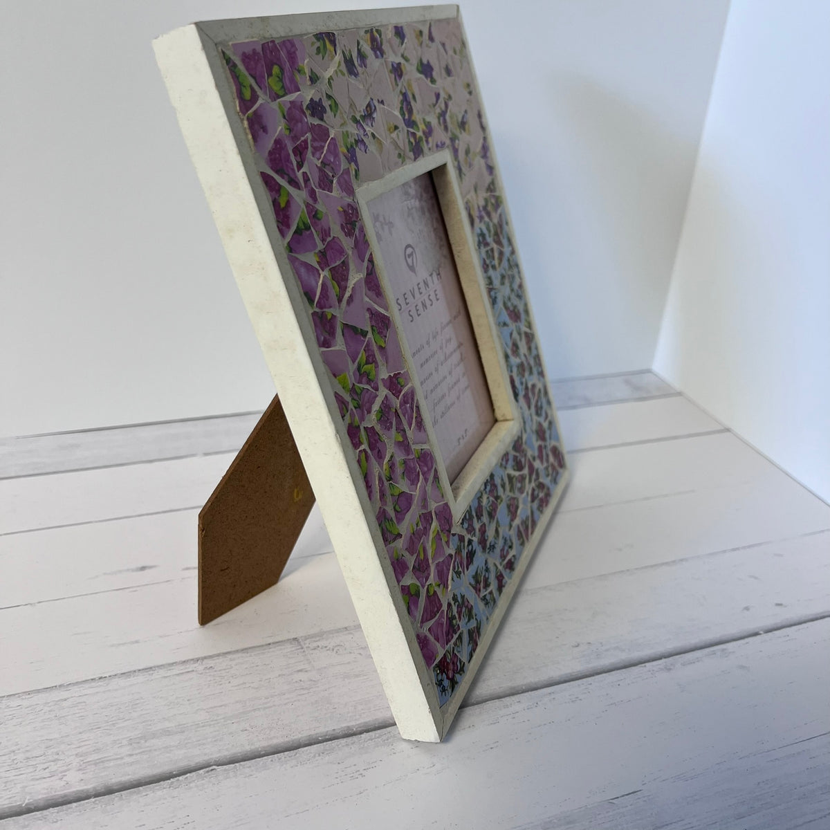 A square mosaic picture frame with a purple wisteria pattern, standing on its backrest on a white wooden surface against a white wall.