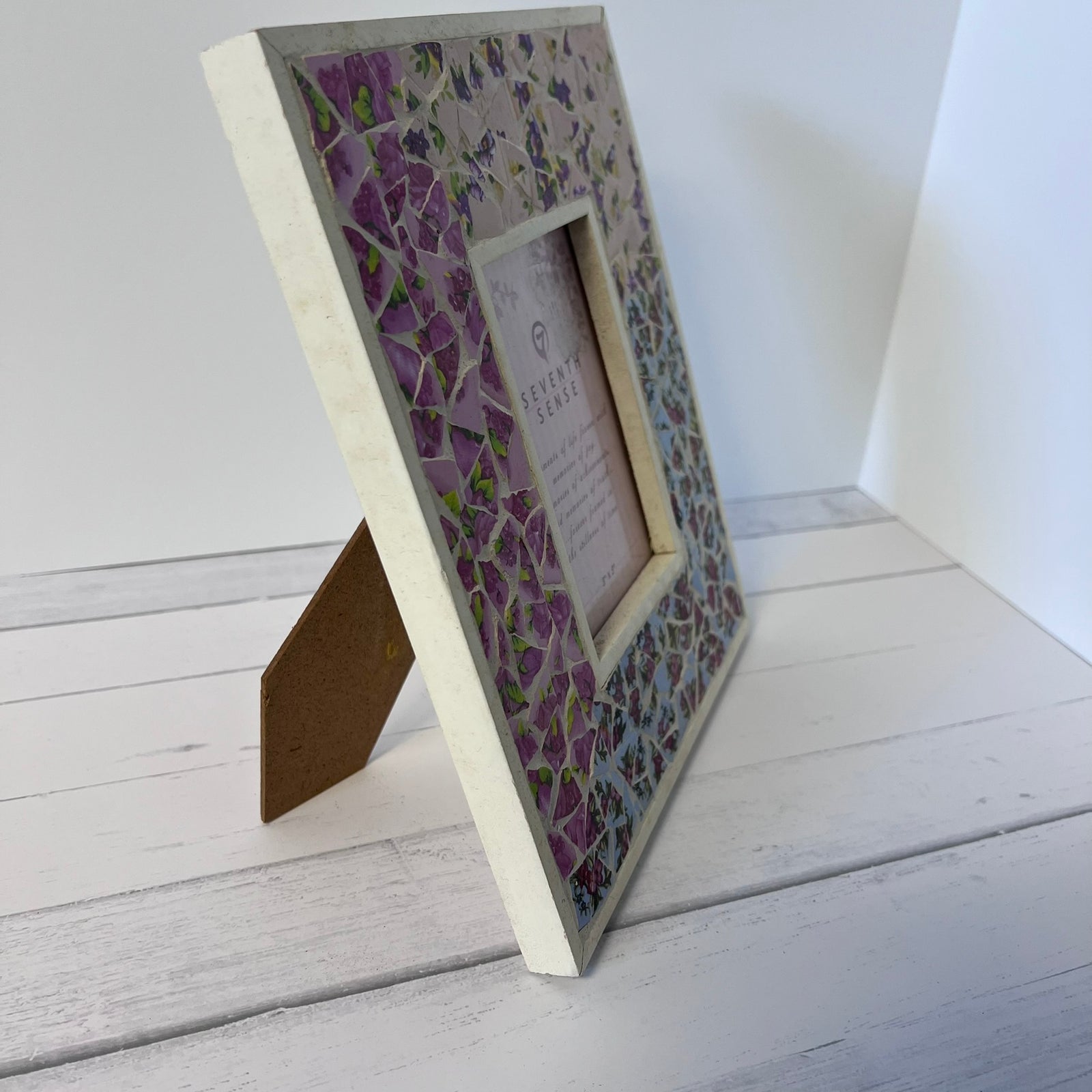 A square mosaic picture frame with a purple wisteria pattern, standing on its backrest on a white wooden surface against a white wall.