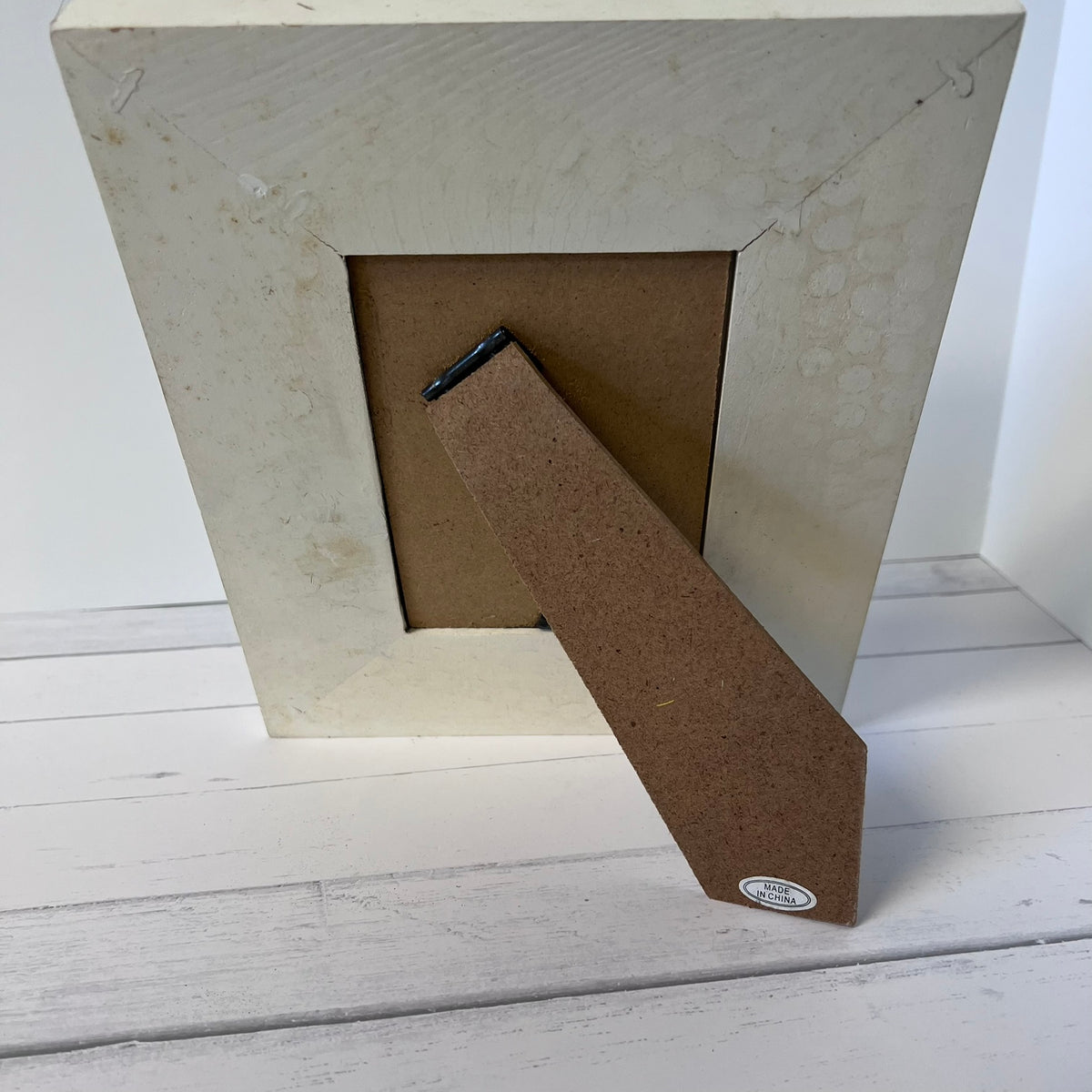 A square picture frame with a textured off-white/cream outer border and a brown cardboard backing, shown standing on a white wooden surface.