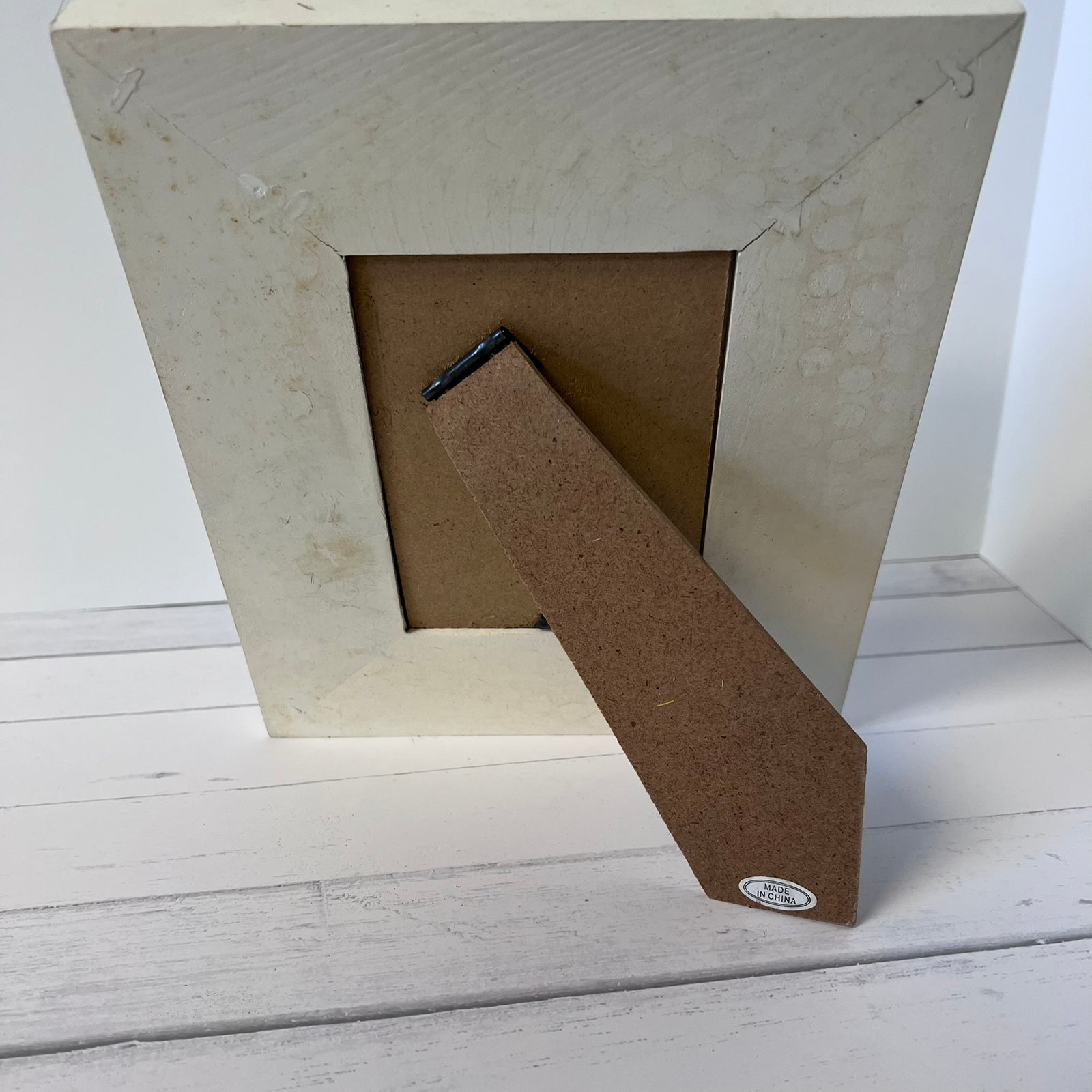 A square picture frame with a textured off-white/cream outer border and a brown cardboard backing, shown standing on a white wooden surface.