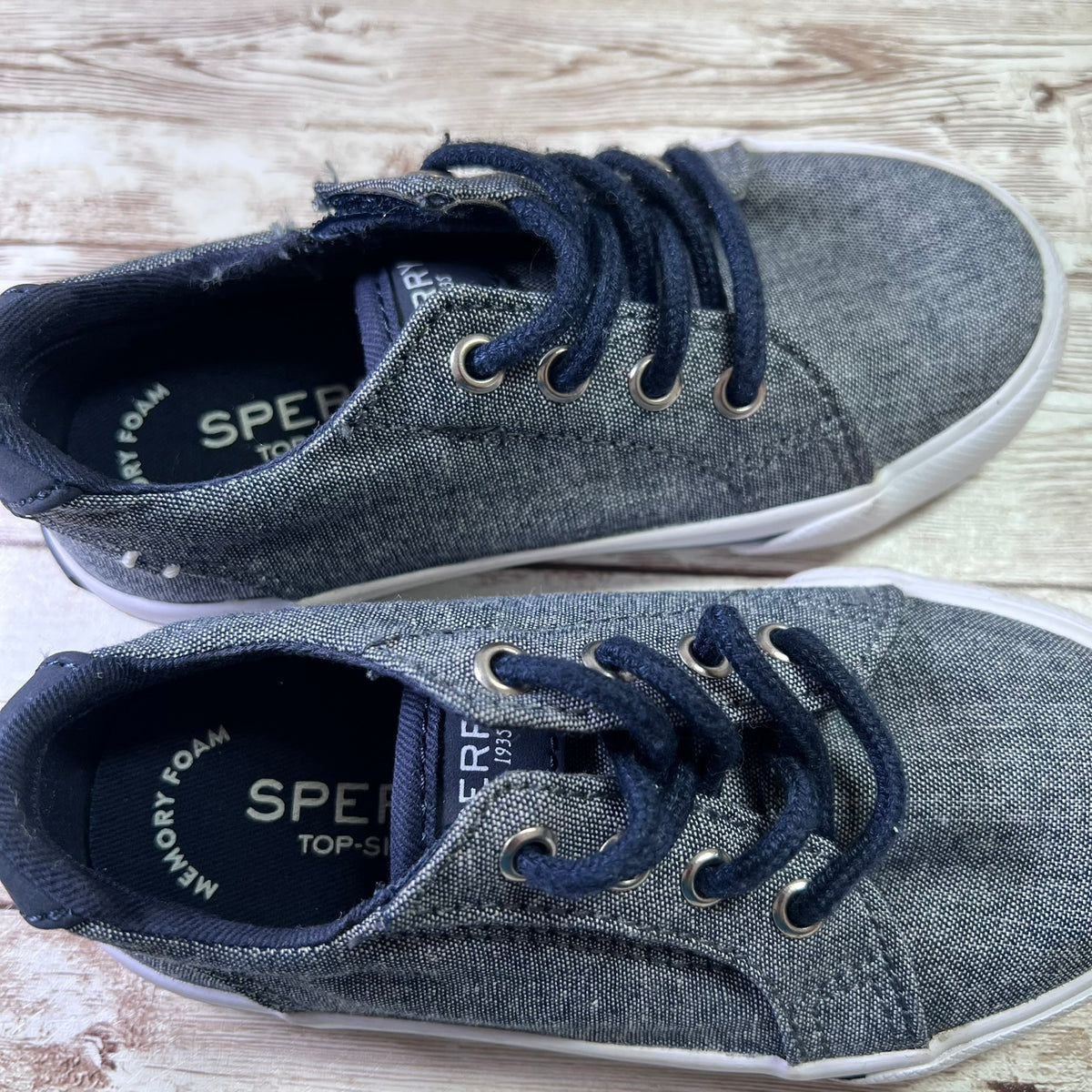 A pair of children's low-top sneakers in a dark gray canvas-like material with navy blue laces and white soles, displayed on a light wood surface. The brand name 'SPECFANTASY' is visible on the inner lining.