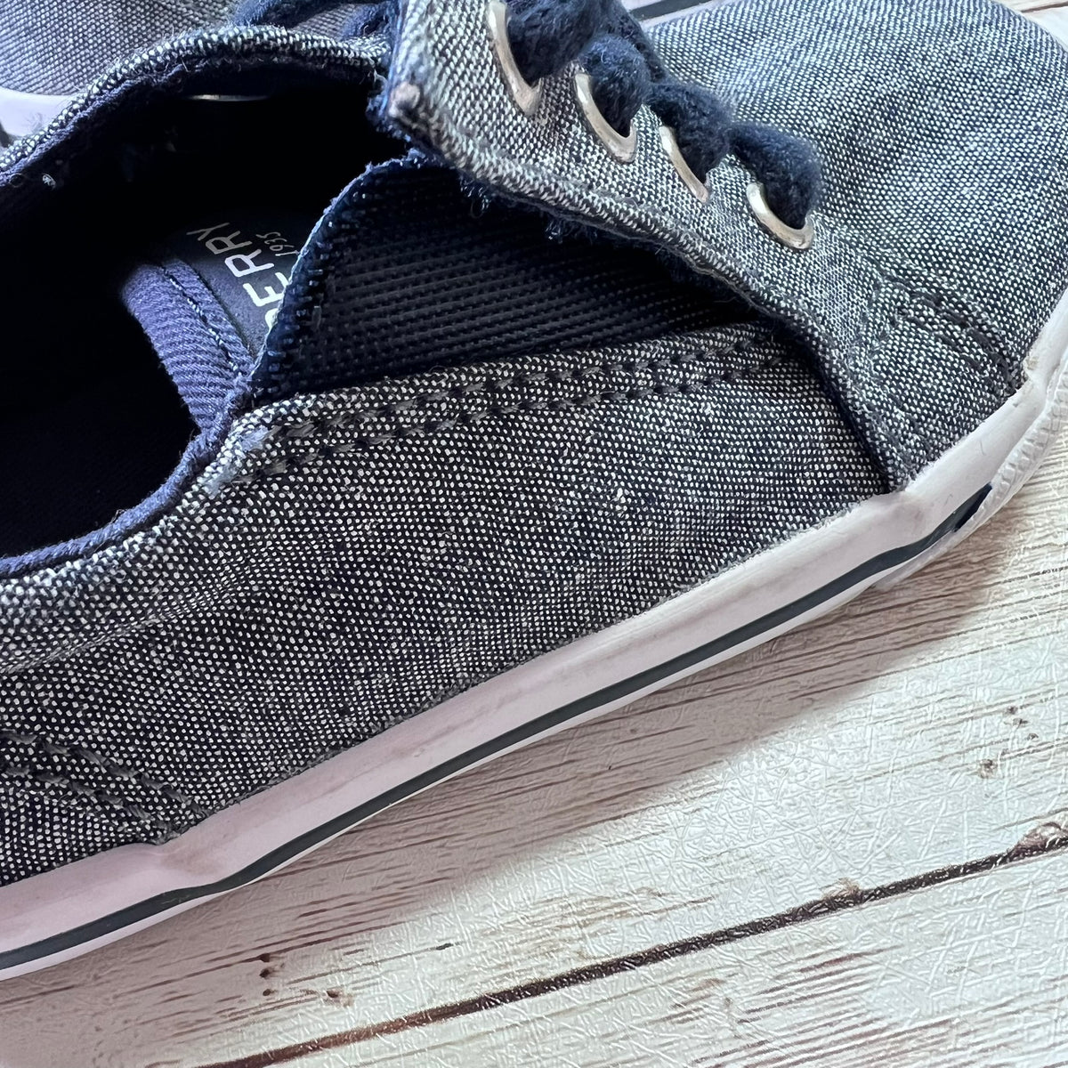 A close-up of a canvas slip-on sneaker in a washed denim blue color, featuring dark navy laces and a white rubber sole, resting on a light-colored wooden surface.