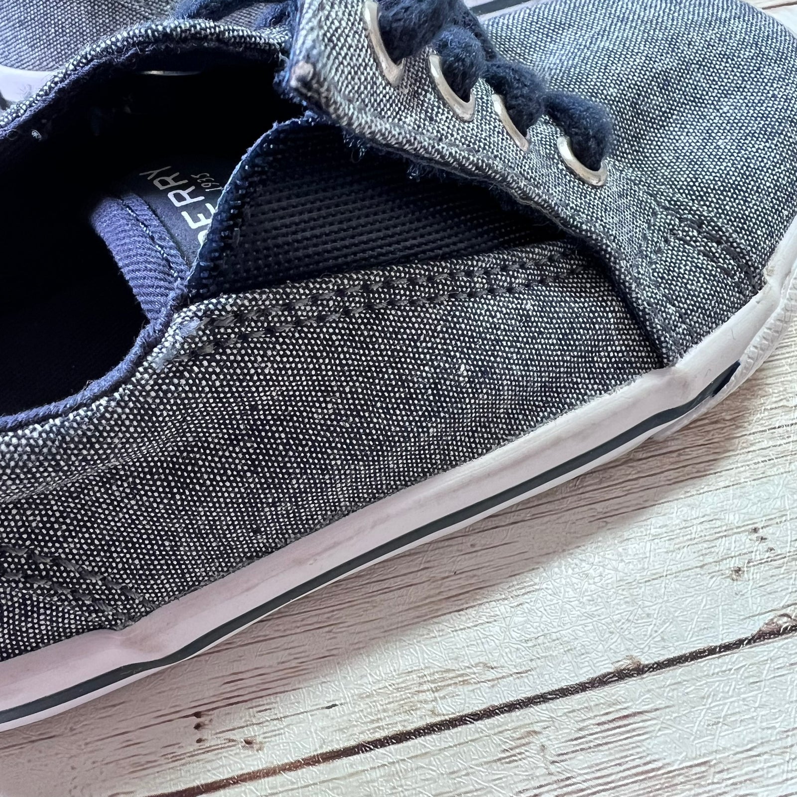 A close-up of a canvas slip-on sneaker in a washed denim blue color, featuring dark navy laces and a white rubber sole, resting on a light-colored wooden surface.