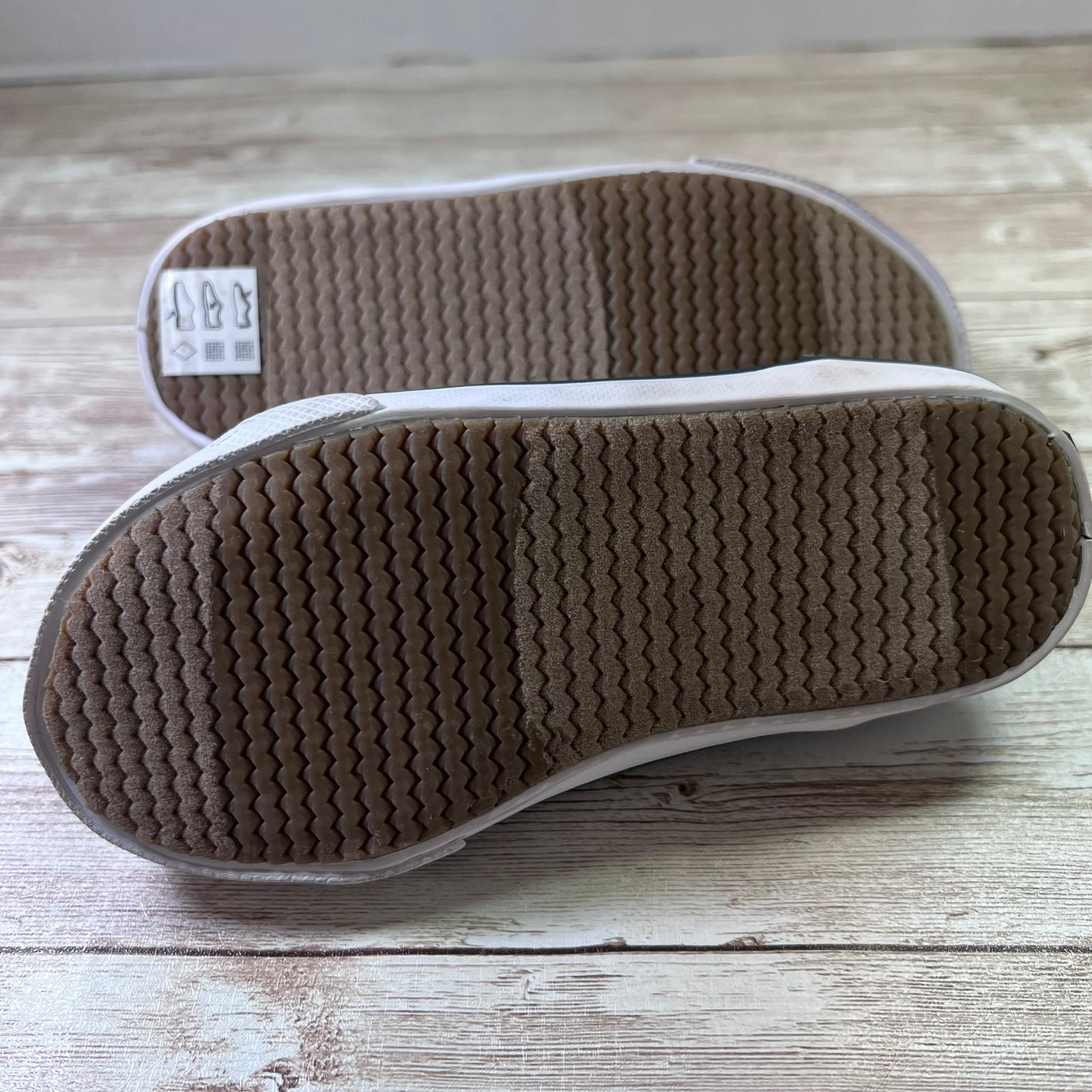 A pair of white slip-on shoes shown from the bottom, displaying their textured brown wavy rubber soles and a size label, placed on a light-colored wooden plank surface.