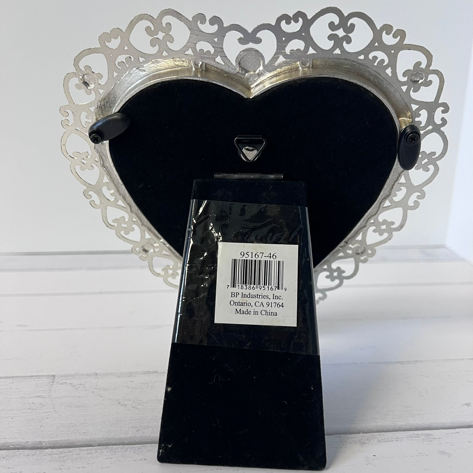 A heart-shaped decorative picture frame with an ornate silver filigree back and a black velvet front, displayed on a black marble base with a product label attached.