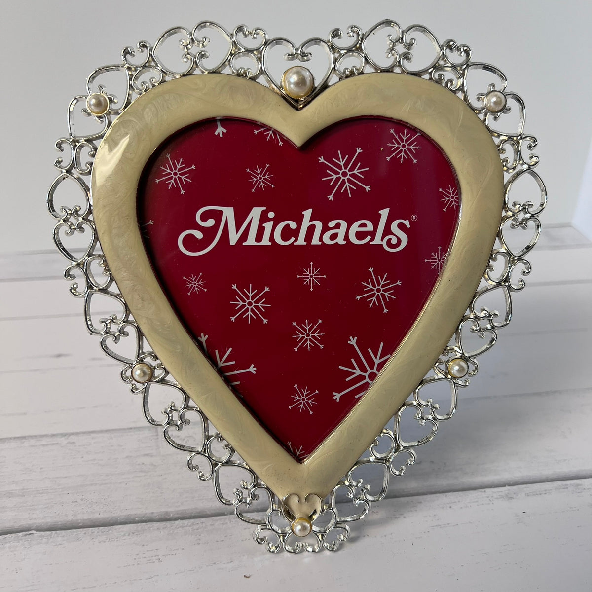 A heart-shaped picture frame with an ornate silver filigree border and a gold-toned inner frame. The center insert is red with white snowflakes and the brand name 'Michaels' in white script.