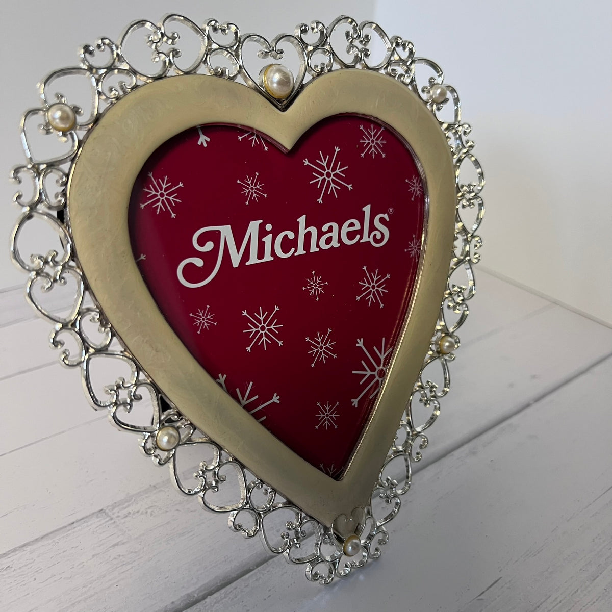 A heart-shaped decorative picture frame with an ornate silver filigree border and pearl accents, featuring a red insert printed with white snowflakes and the name 'Michaels' in white script.