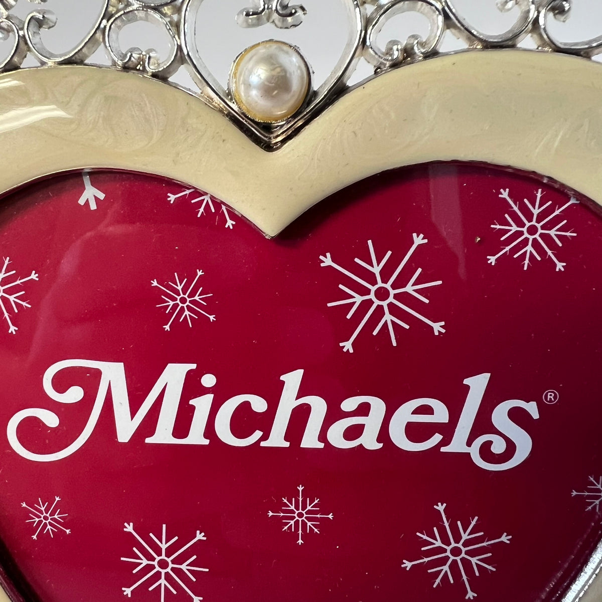 A close-up of a heart-shaped red decorative item with white snowflake motifs and the brand name 'Michaels' in white script. The top edge features a cream-colored border and a silver filigree frame with a pearl-like center.