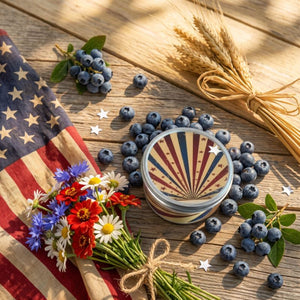 Candle with star design surrounded by blueberries, flowers, and an American flag on a wooden surface.