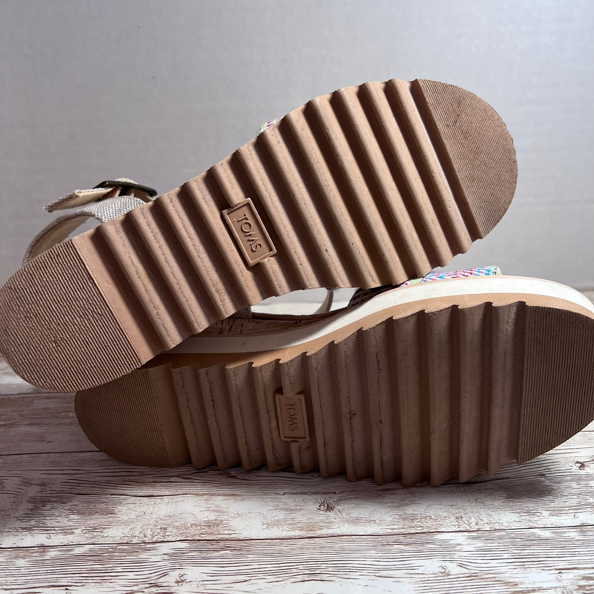 A pair of Toms open-toe sandals shown from the sole side, resting on a rustic wooden surface against a neutral gray background.