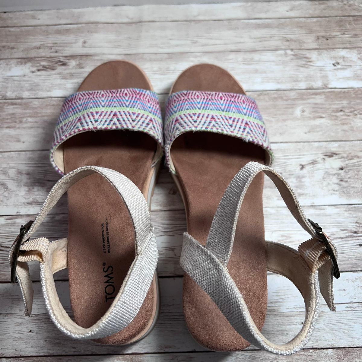 A pair of TOMS women's wedge sandals with beige woven straps and a colorful geometric patterned upper, shown on a light wood plank background.