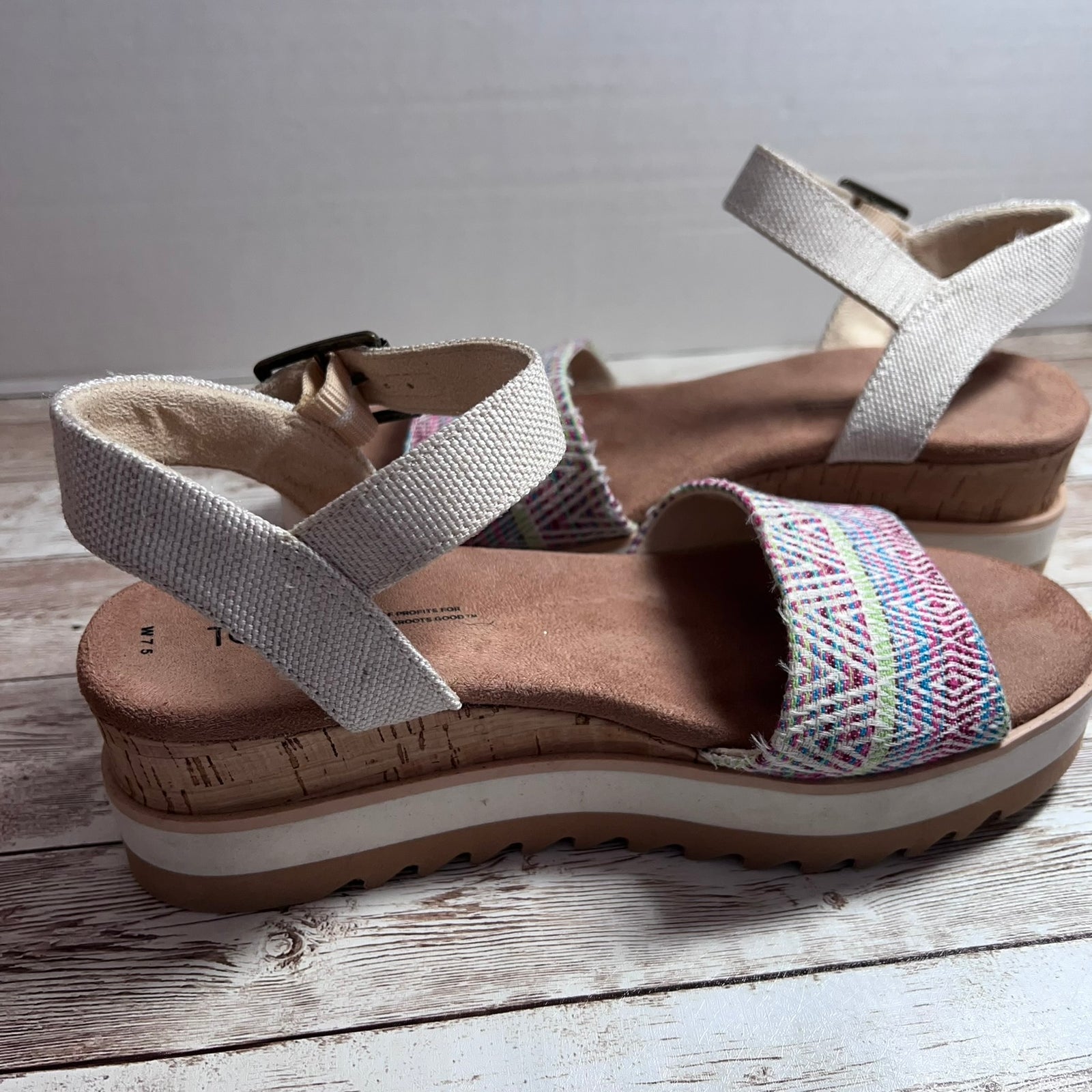 A pair of platform wedge sandals with woven multicolor straps and light beige elastic ankle straps, displayed on a rustic white-washed wooden surface.
