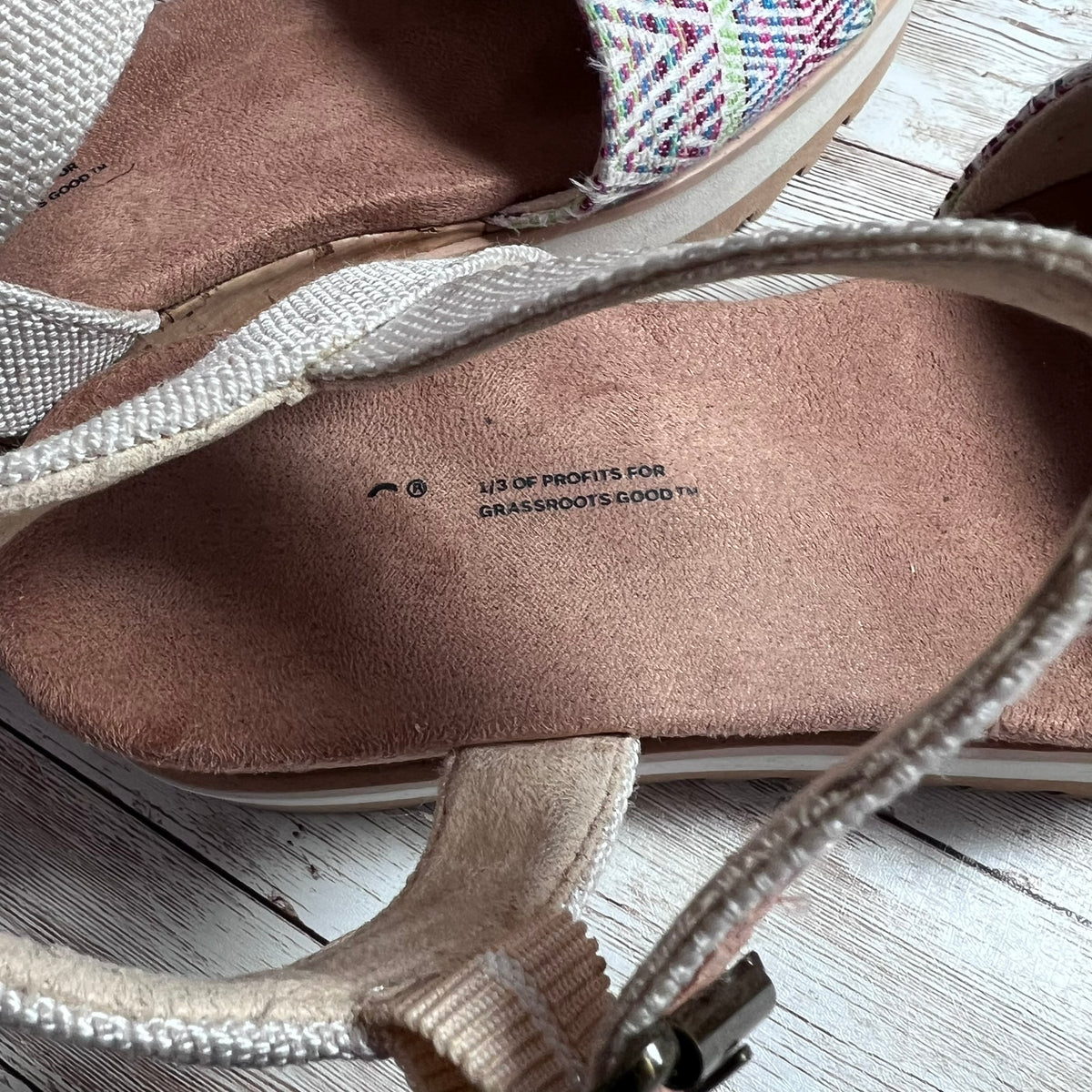 Close-up of the interior of a wedge sandal showing a brown suede footbed with printed text reading '1/3 OF PROFITS FOR GRASSROOTS-GOOD™', a woven multicolor side panel, and beige straps on a whitewashed wood surface.