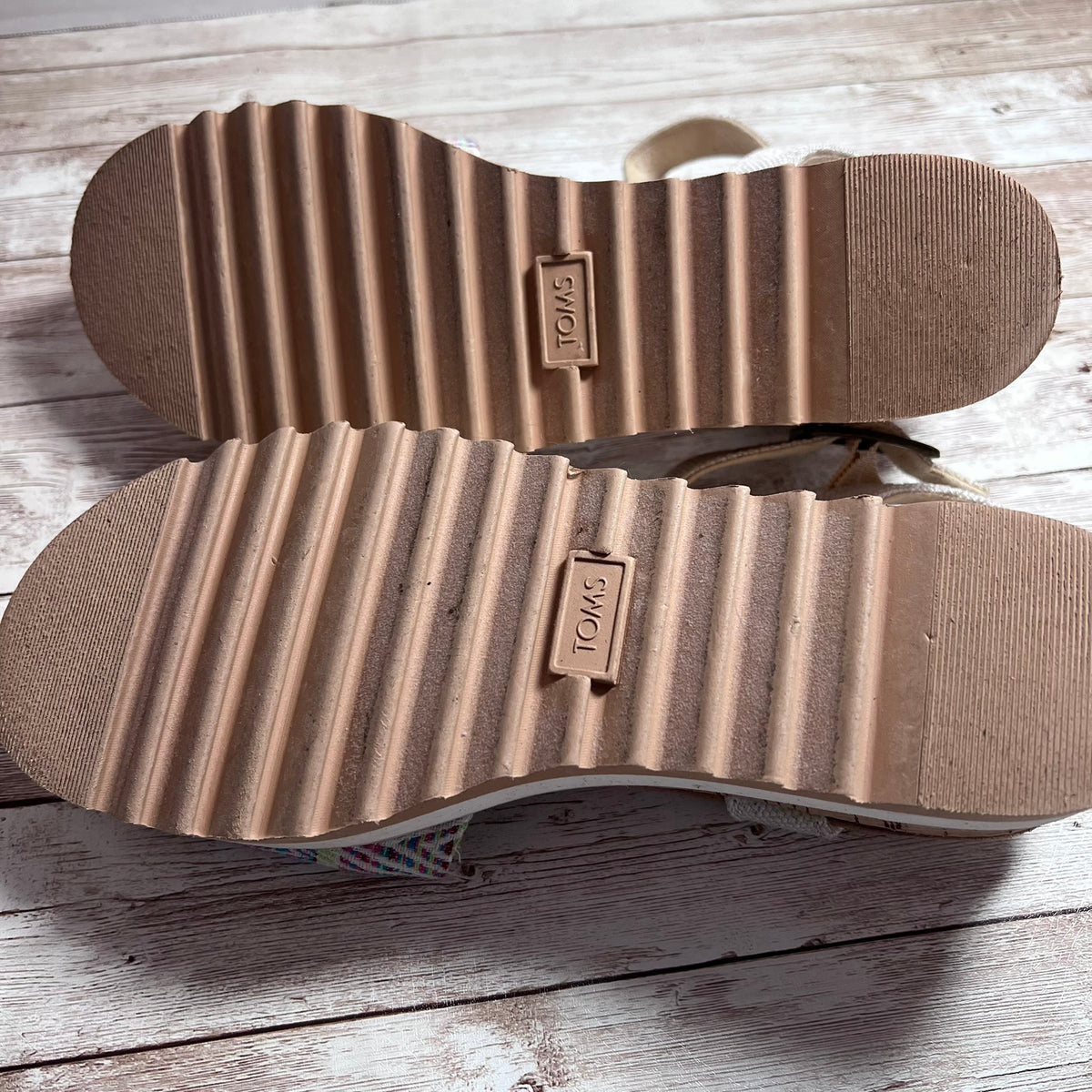 The soles of a pair of TOMS sandals, showing a ribbed tan rubber tread with a small embossed brand tag reading 'TOMS' in the center, photographed on a light rustic wooden surface.