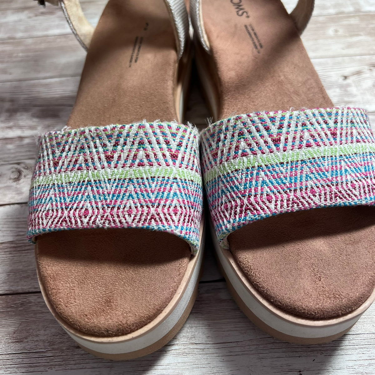 A pair of wedge sandals with tan suede-like footbeds and thick platform soles, featuring wide woven upper straps in pink, blue, green, and white. The sandals are photographed on a light gray wood plank surface.