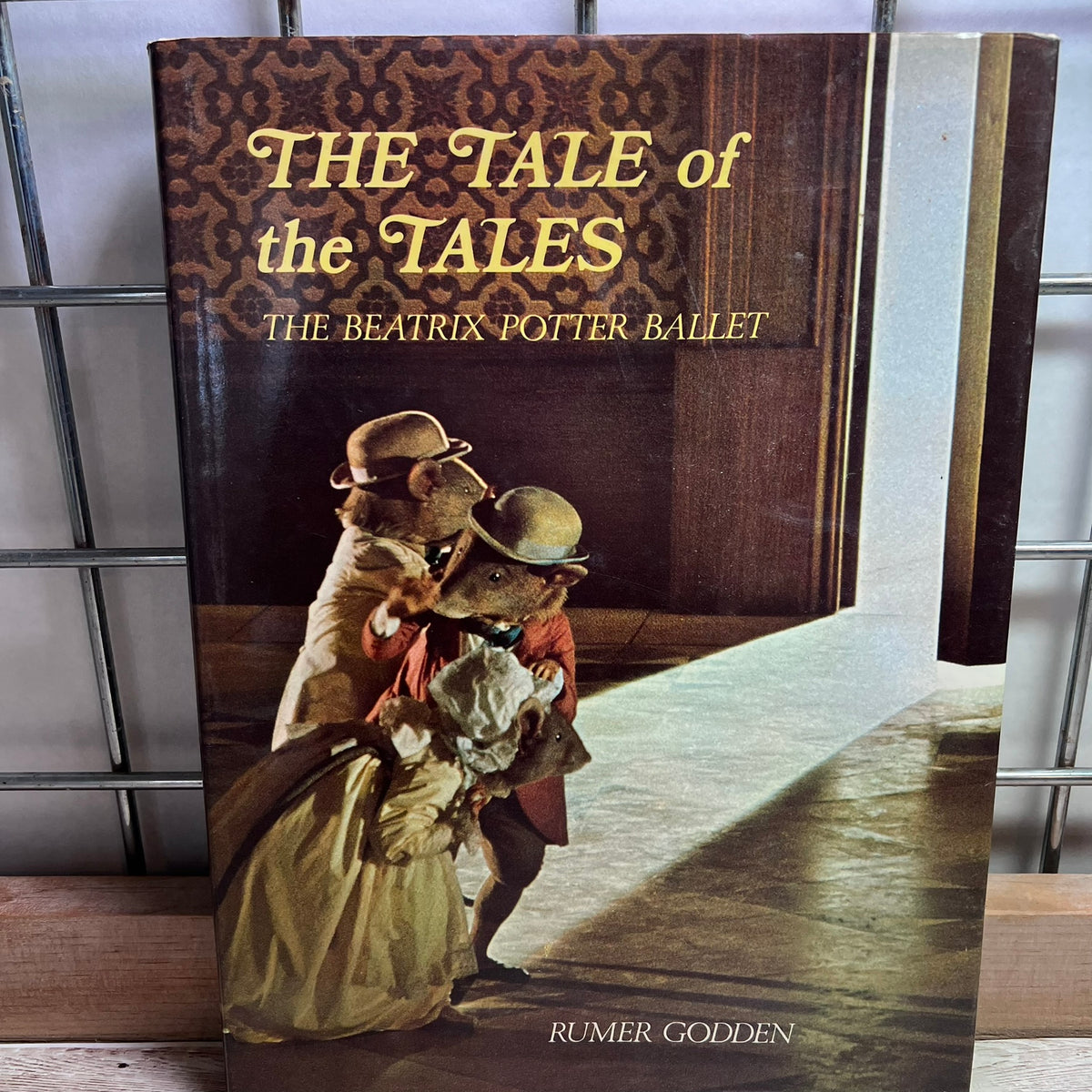 A hardcover book titled "The Tale of the Tales: The Beatrix Potter Ballet" by Rumer Godden, standing upright on a shelf. The cover features vintage-style illustrations of foxes in period costumes under a patterned brown header, with a warm spotlight effect on the floor.