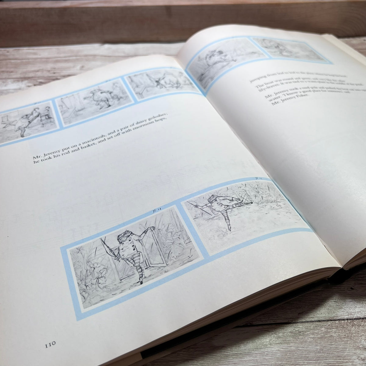 An open book lying on a rustic wooden surface, showing printed pages with black line drawings and text framed by pale blue borders.