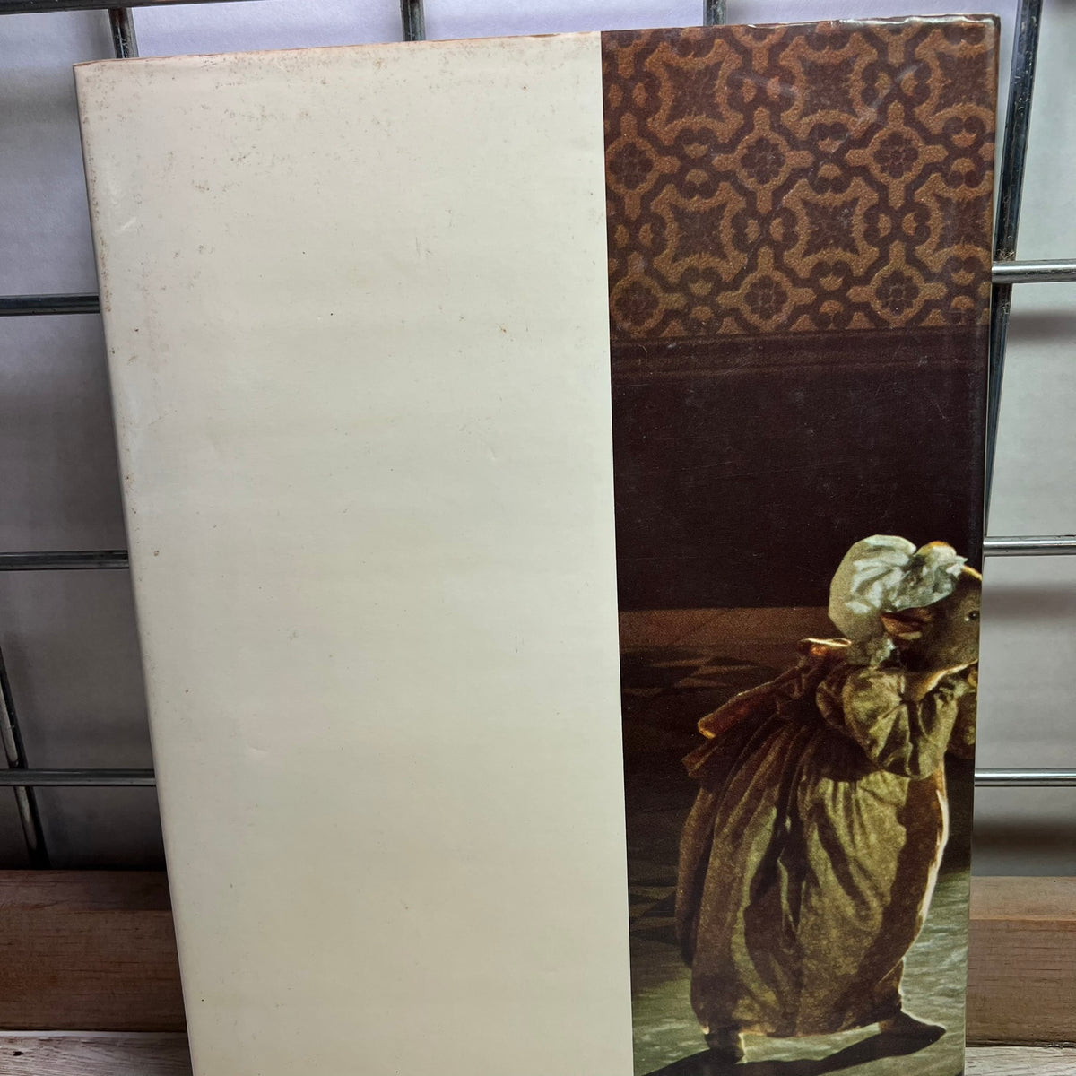 A vintage art print or book cover standing upright on a shelf. The left half is a blank warm off-white/cream area, while the right half shows a classical painting of a woman in a long dress carrying a child, with a brown patterned top section above.