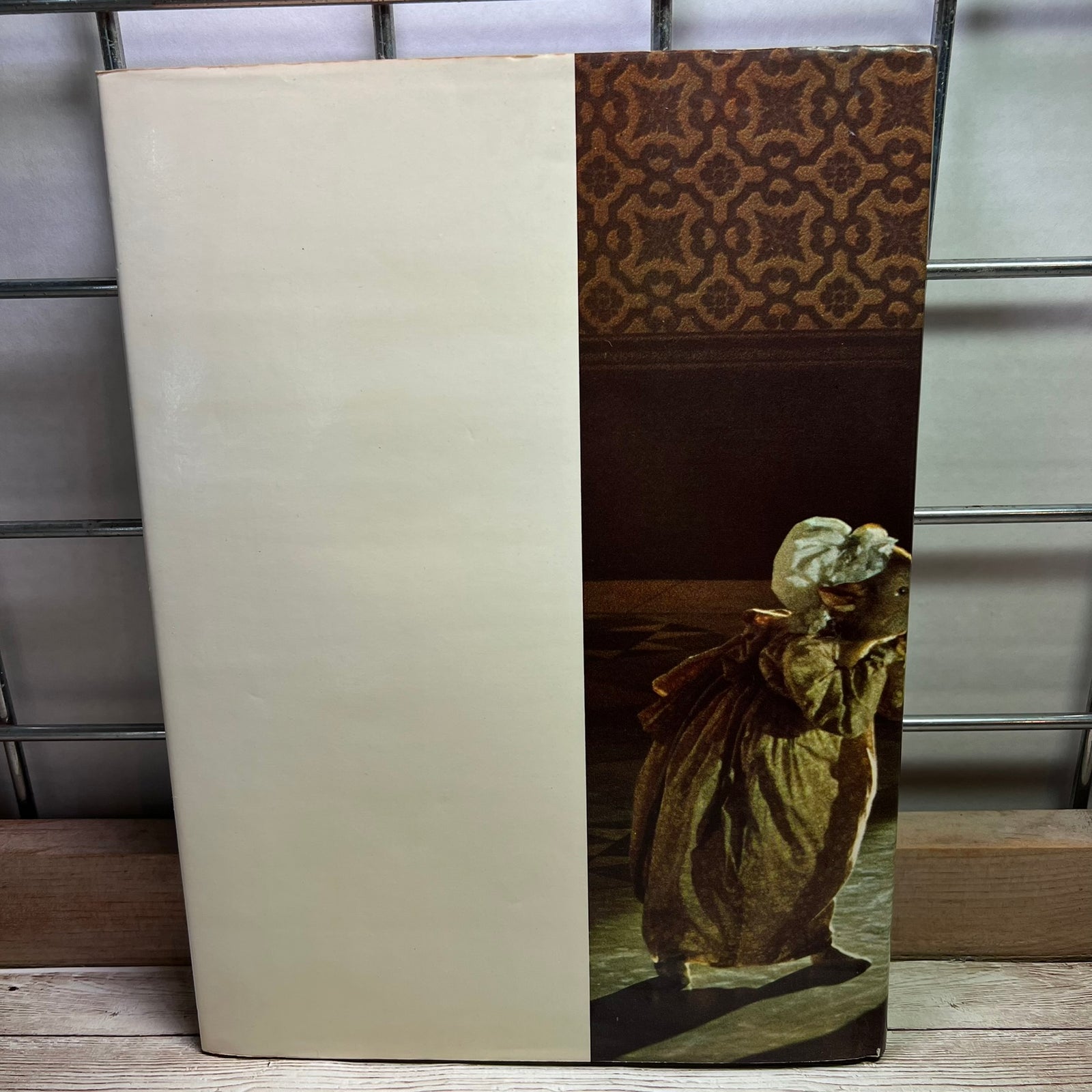 A vintage art book or portfolio standing upright on a wooden surface. The cover is split vertically: the left half is a plain warm cream/beige, and the right half features a dark classical painting of a woman in a long dress, with a brown patterned border at the top.