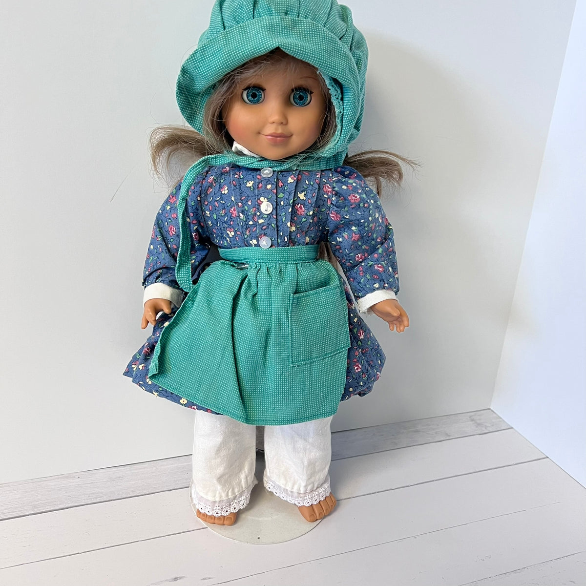 A doll with brown hair and blue eyes wearing a teal green bonnet, a blue floral long-sleeve top, a solid green apron with a front pocket, and white pants, standing against a white wall and light wood floor.