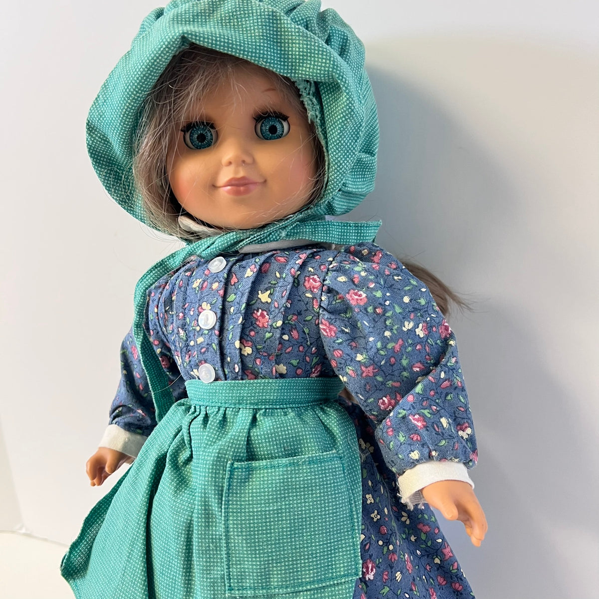 A doll with blue eyes and gray hair wearing a teal bonnet and a blue floral dress with a teal apron, photographed against a plain light background.