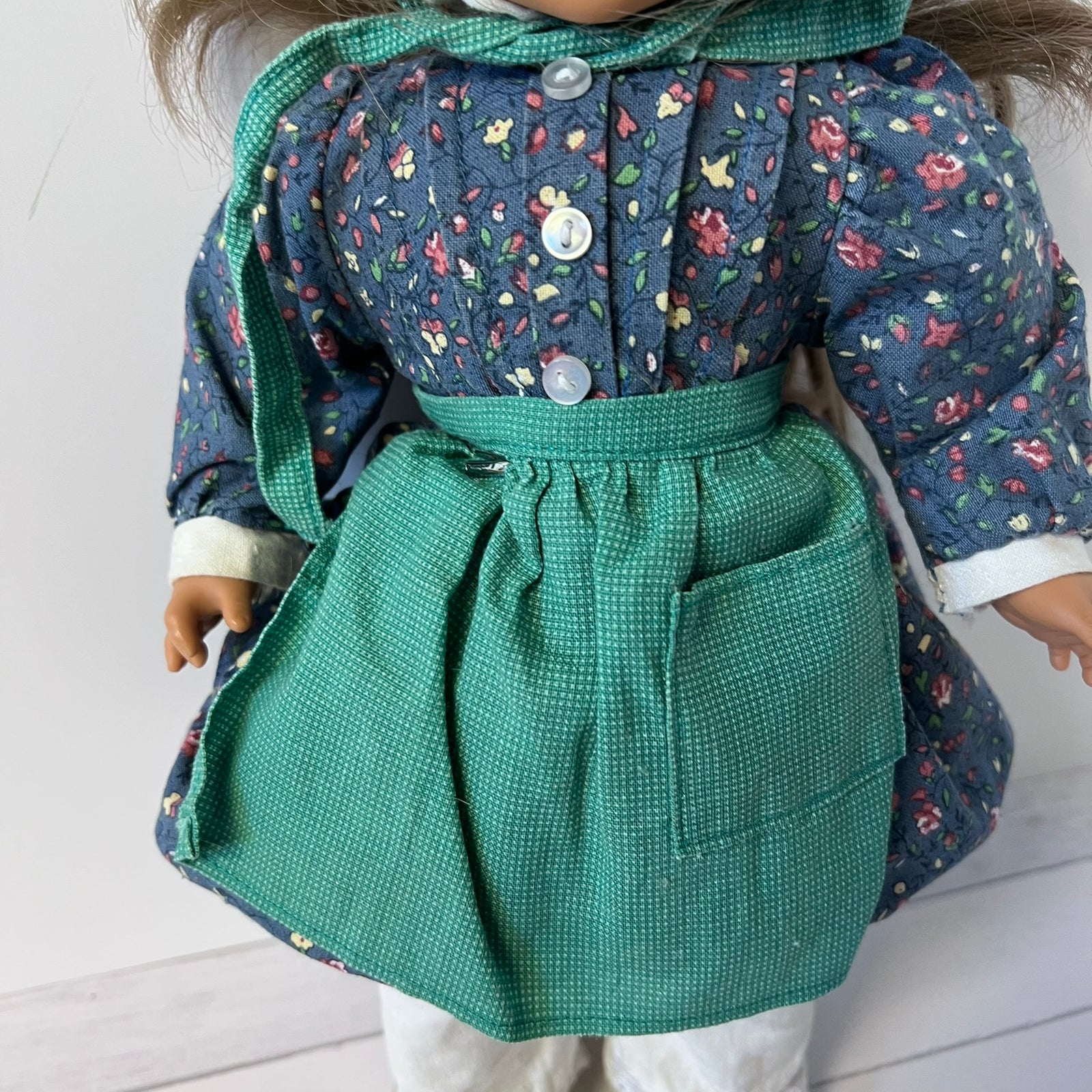 A close-up of a doll wearing a floral print jacket with a green hood and a textured green apron-style skirt with a front pocket.