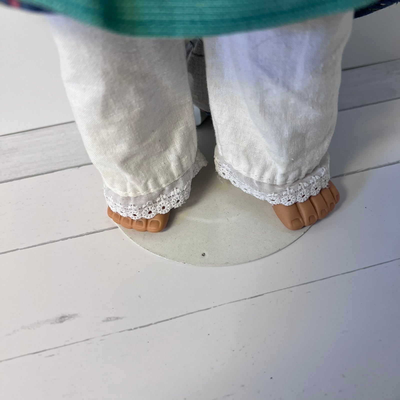 Close-up of a doll's lower legs wearing off-white linen pants with white lace trim and brown plastic feet, standing on a round platform over a white wood plank floor.