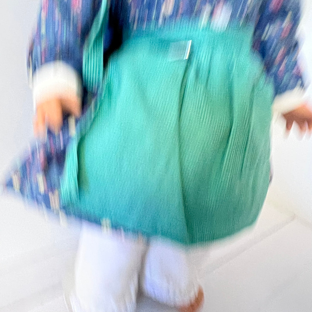 A doll or toy figure wearing a teal corduroy-style pinafore dress over a blue patterned jacket and white pants, photographed against a plain white background. The image is heavily blurred and motion-like, making details soft and abstract.