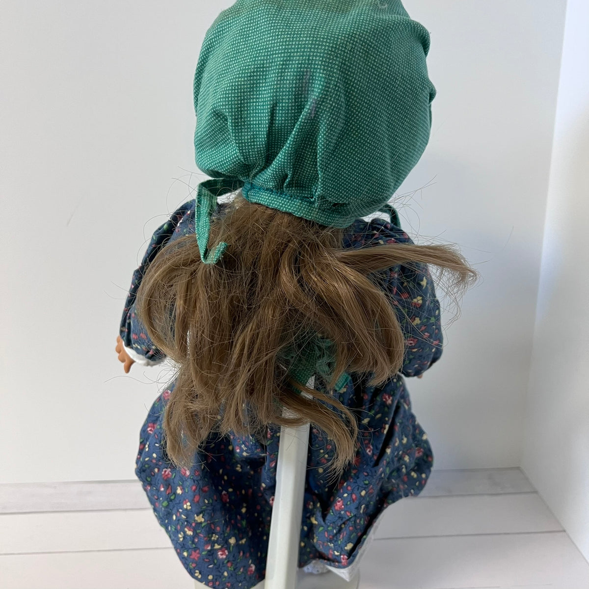 A doll viewed from behind wearing a textured green bonnet and a dark blue floral dress, sitting on a white chair against a white wall.