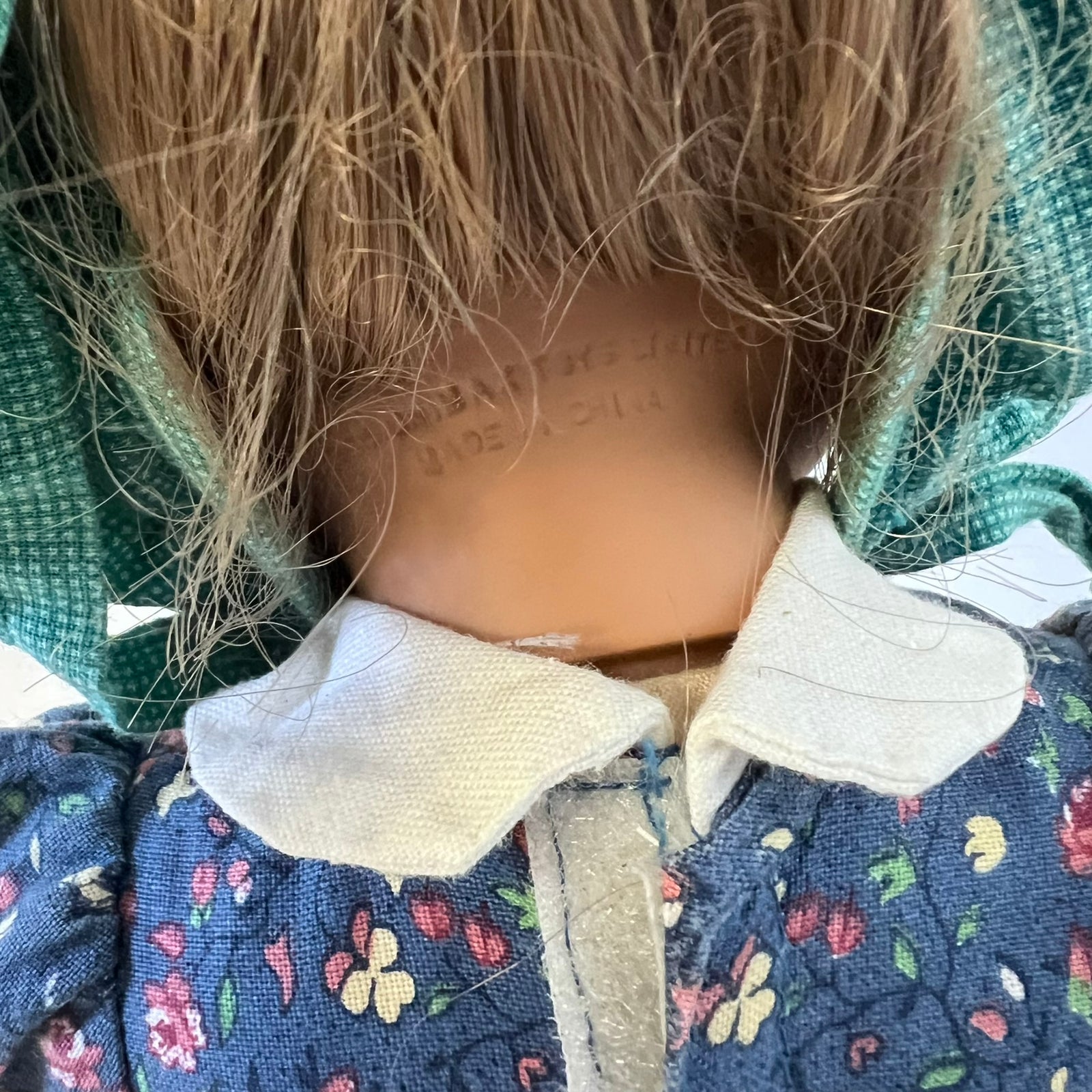 Close-up back view of a doll or toy figure wearing a floral blue dress with a cream fabric collar, brown hair, and a green patterned head covering.