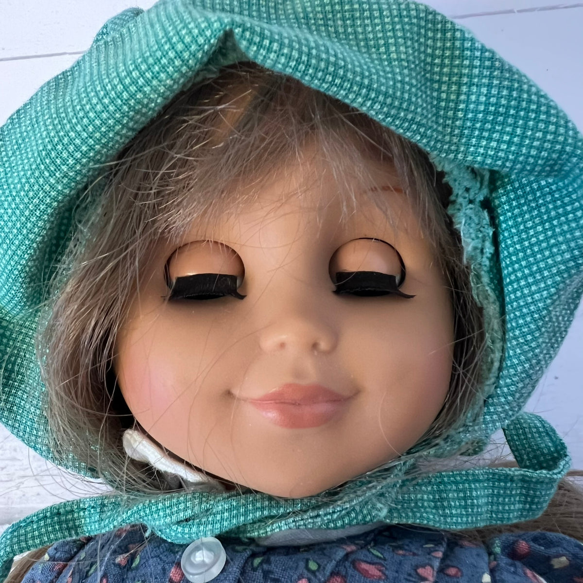 A close-up of a doll's face wearing a green patterned bonnet and a blue floral dress against a white wooden background.