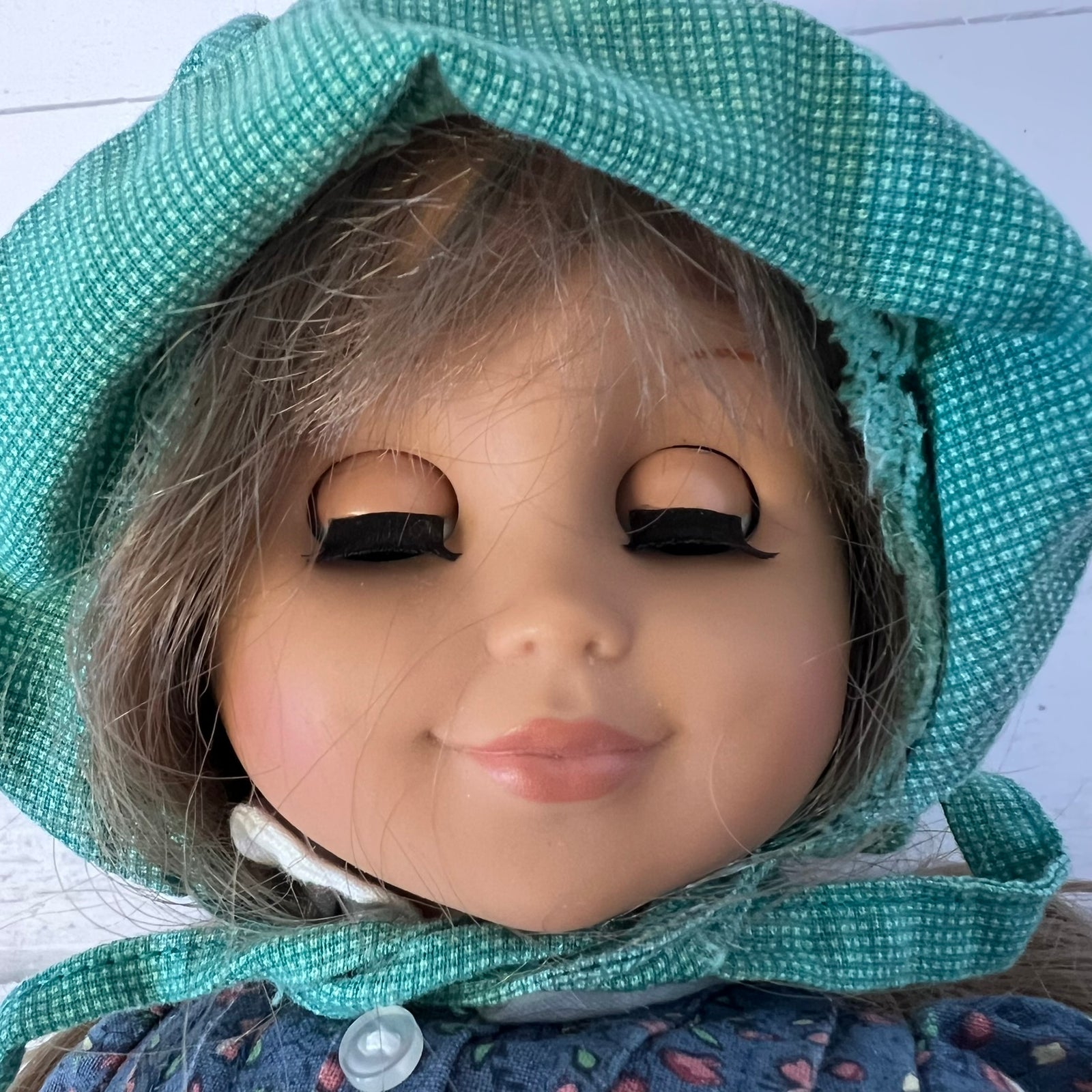 A close-up of a doll's face wearing a green patterned bonnet and a blue floral dress against a white wooden background.