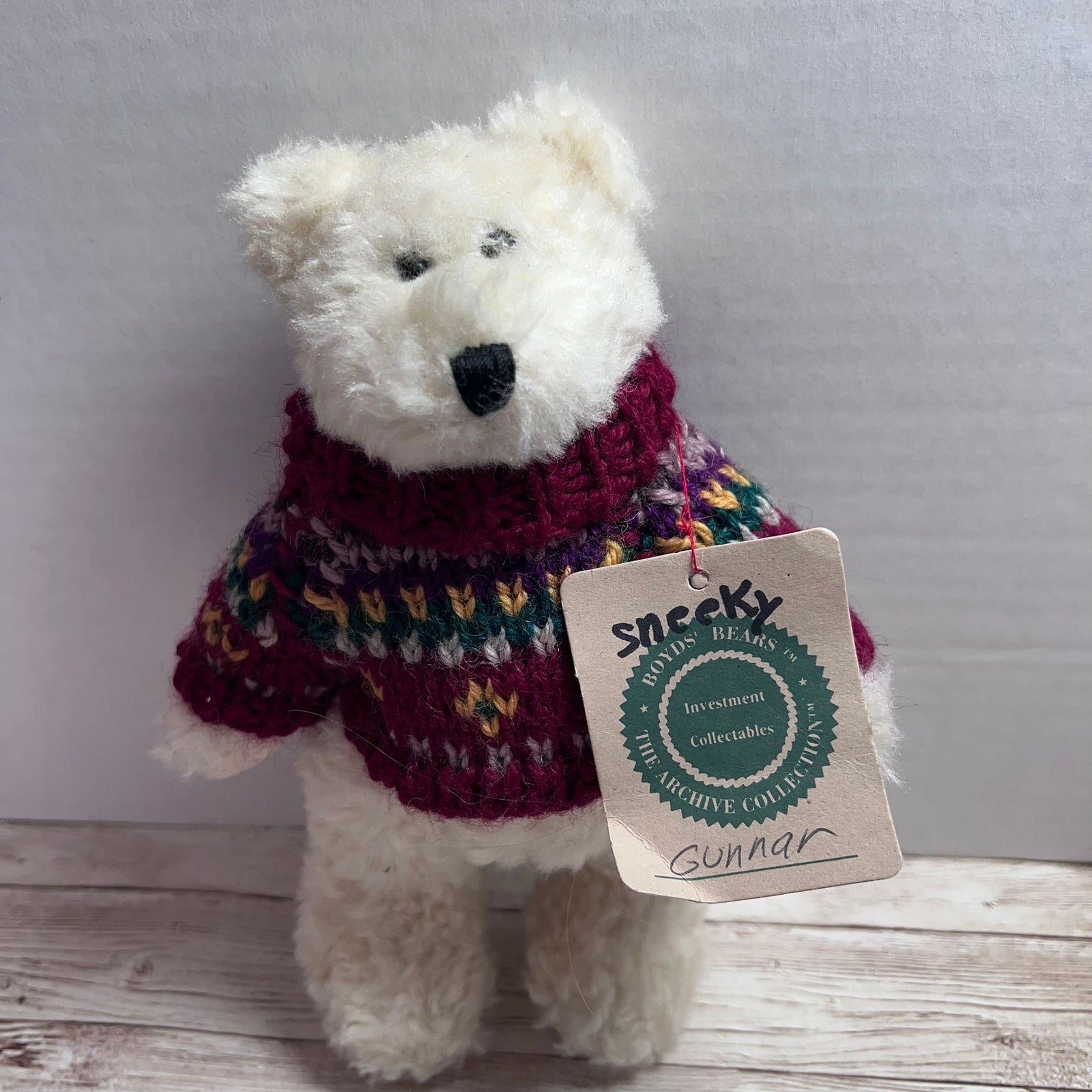A small white plush teddy bear wearing a knitted multicolor sweater, standing against a light gray wall on a pale wood surface. A paper hangtag is attached and reads 'Sneeky Handmade Collectibles' with the name 'Gunnar' written at the bottom.