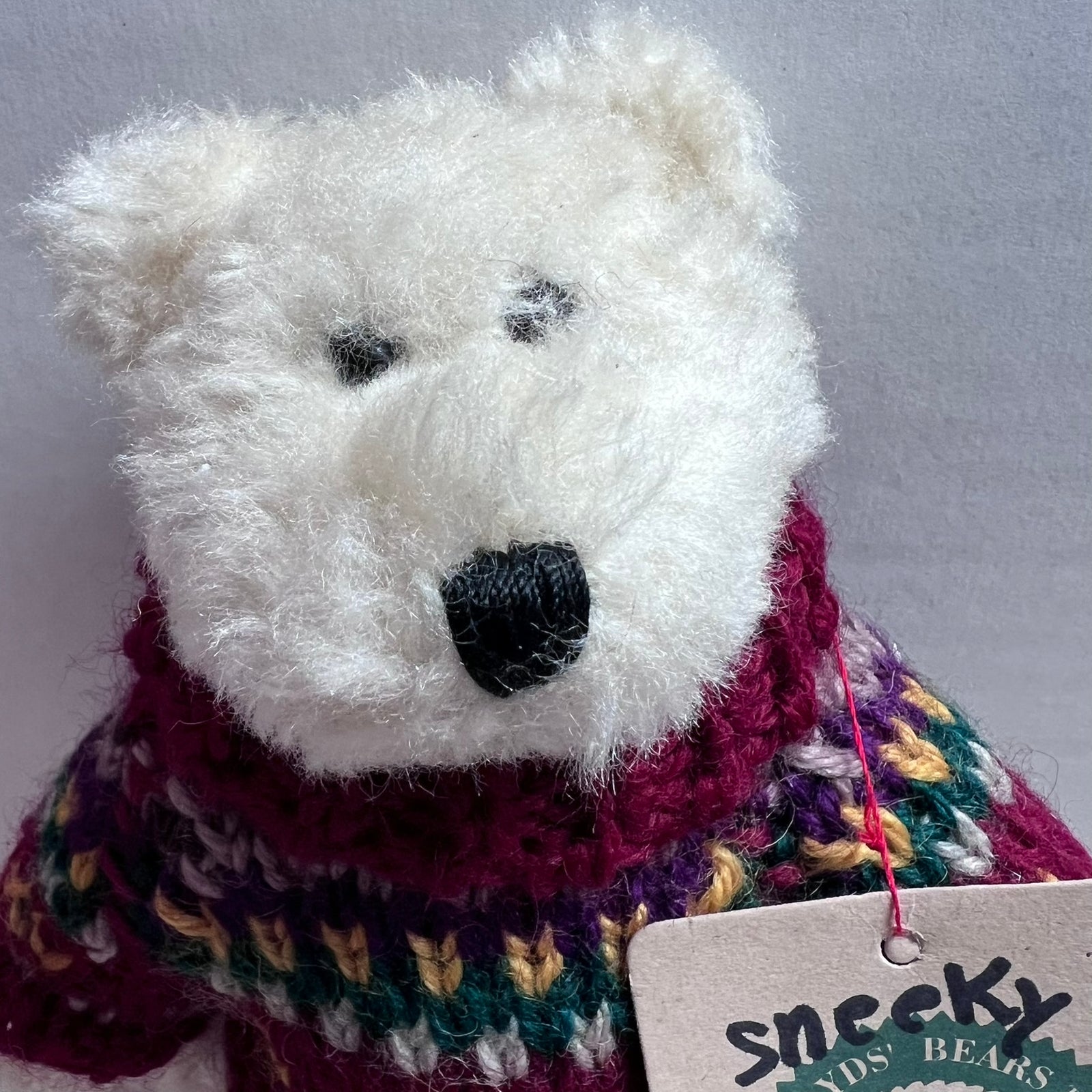 A close-up of a white plush teddy bear wearing a chunky knit sweater in burgundy, purple, green, and yellow, with a paper tag reading 'snickz kids bears' attached.