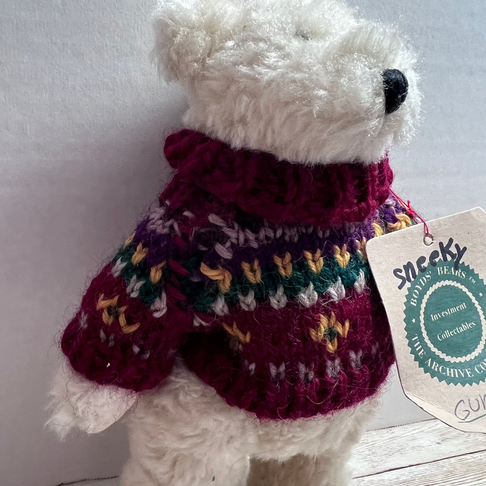 A white plush teddy bear wearing a hand-crocheted multicolor sweater with a deep burgundy neck and body, featuring purple, green, yellow, and white patterns. A paper hangtag from 'Snickky Toy Archives' is attached to the side.