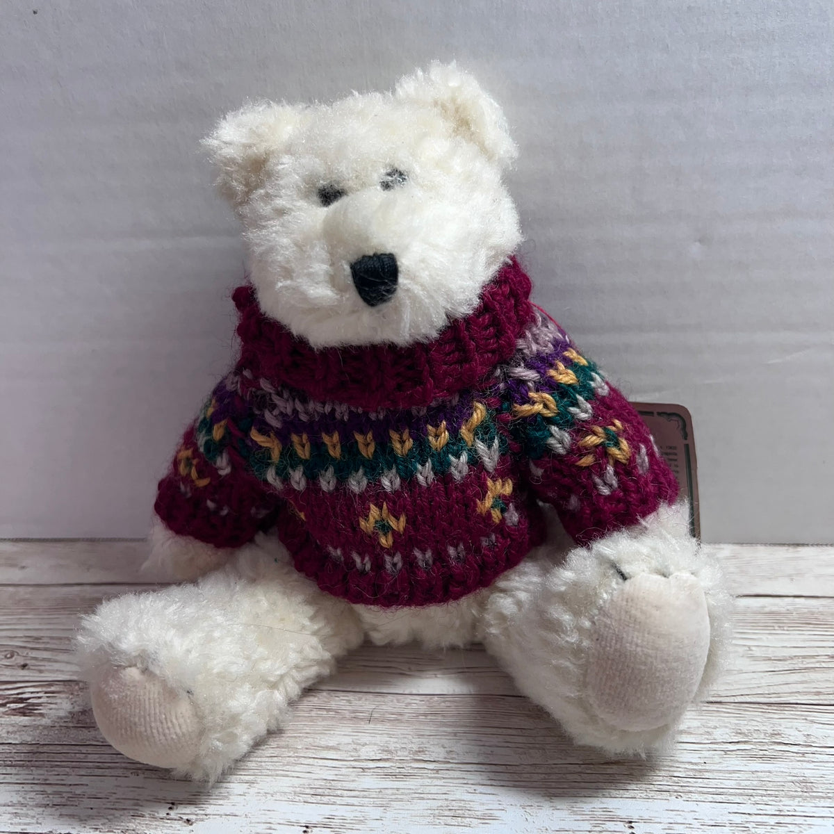 A white plush teddy bear wearing a knitted multicolor sweater with a burgundy base and green, yellow, and white Fair Isle style pattern, sitting on a rustic white wooden surface.