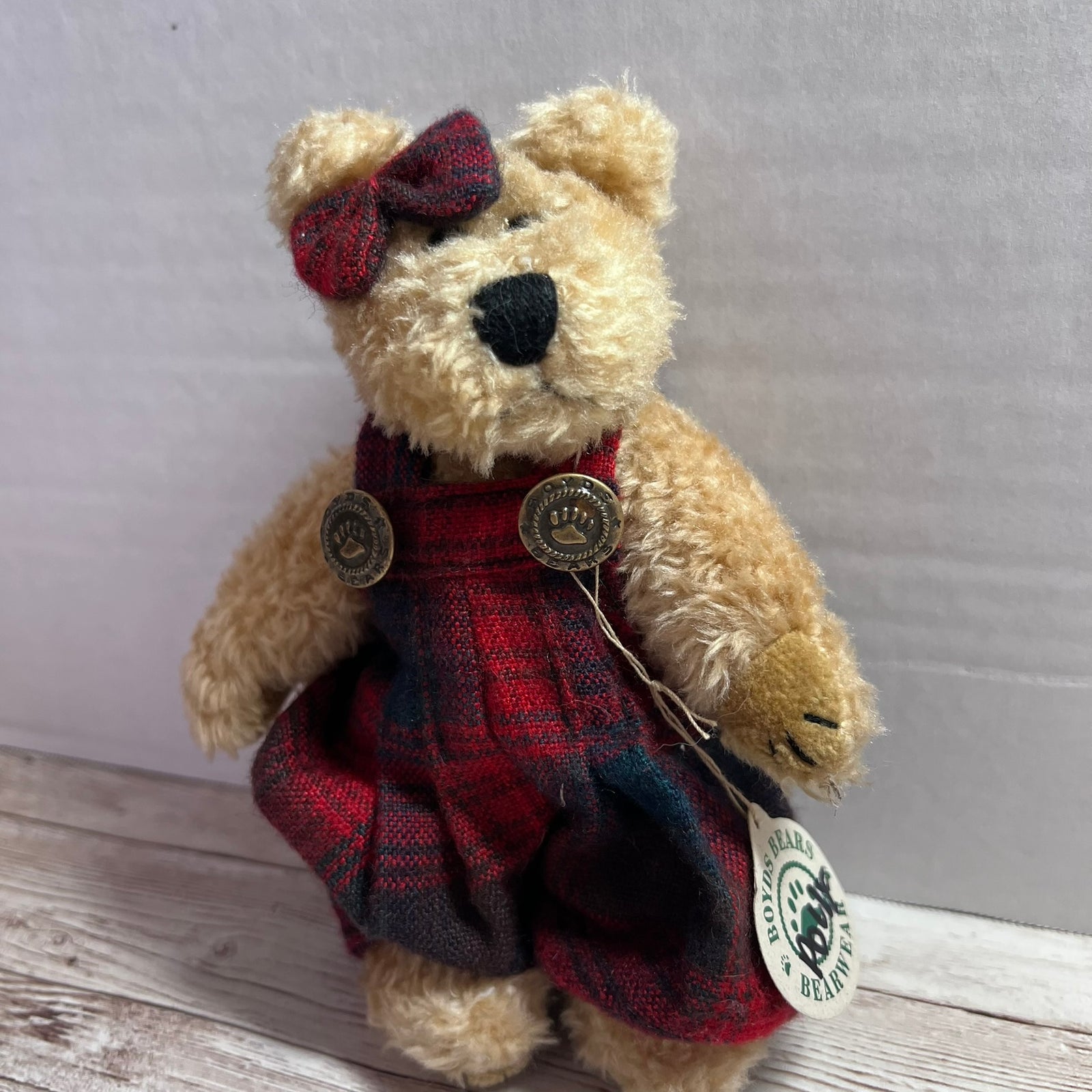A plush teddy bear with light tan fur wearing red and black plaid overalls, a matching plaid bow headband, and a hangtag that reads 'Handmade Bear'.