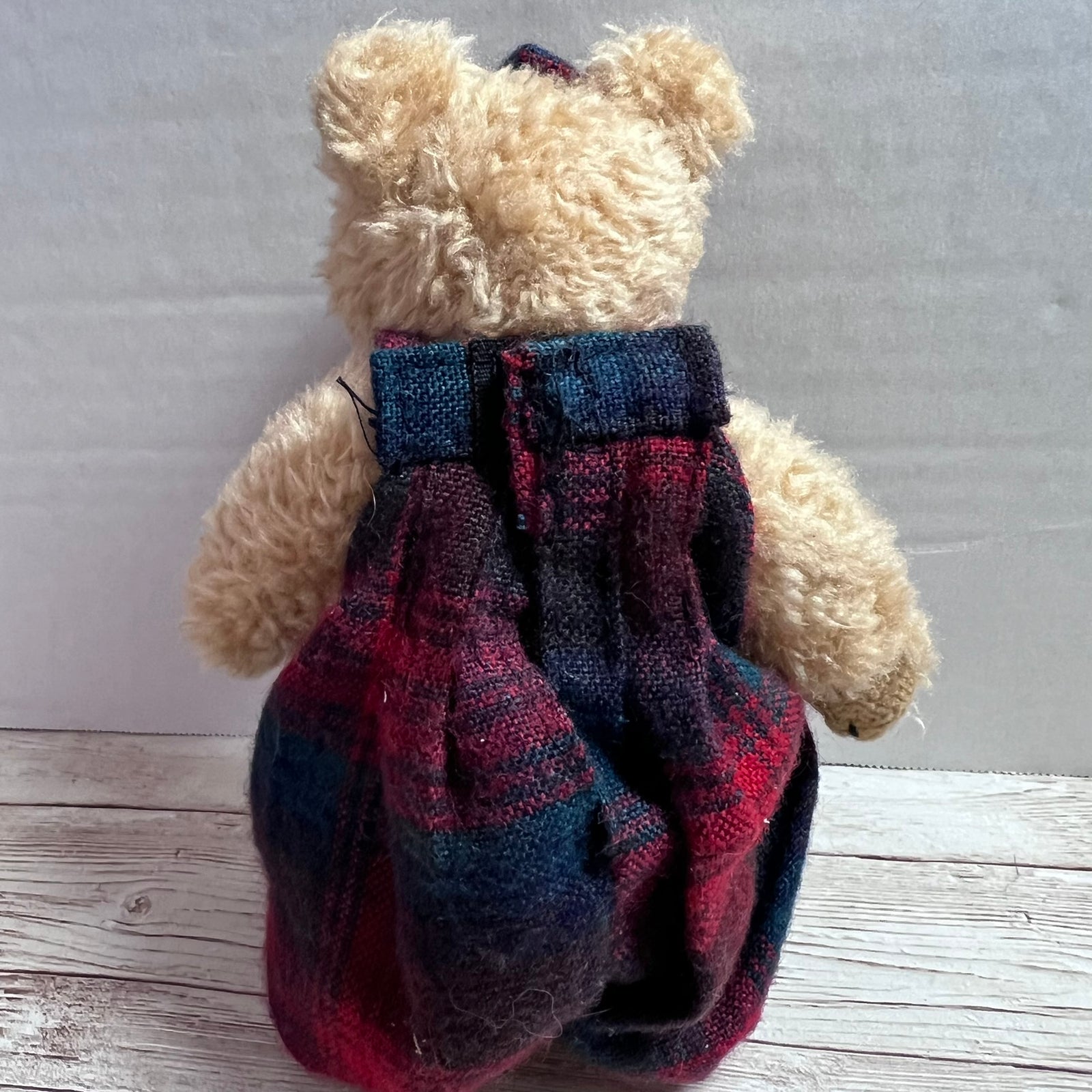 A small beige plush teddy bear viewed from the back, wearing a red, navy, and black plaid dress over a dark top, standing on a light wood surface against a plain gray background.
