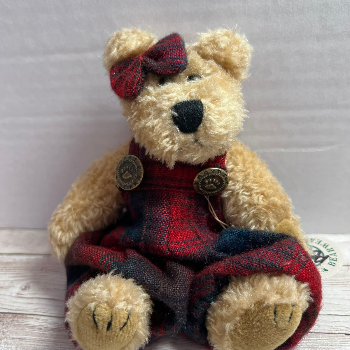 A plush teddy bear with cream-colored fur wearing red and navy tartan plaid overalls and a matching bow on its head, sitting on a light wood surface against a gray background.