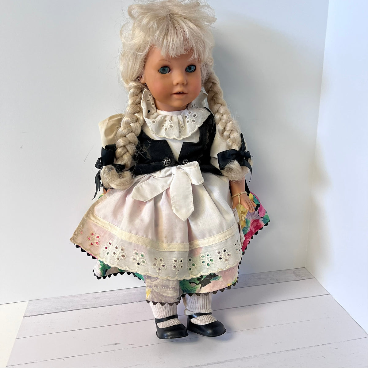 A vintage-style porcelain doll with long blonde pigtails, wearing a black and white dress with a large white bow, standing against a plain white wall.
