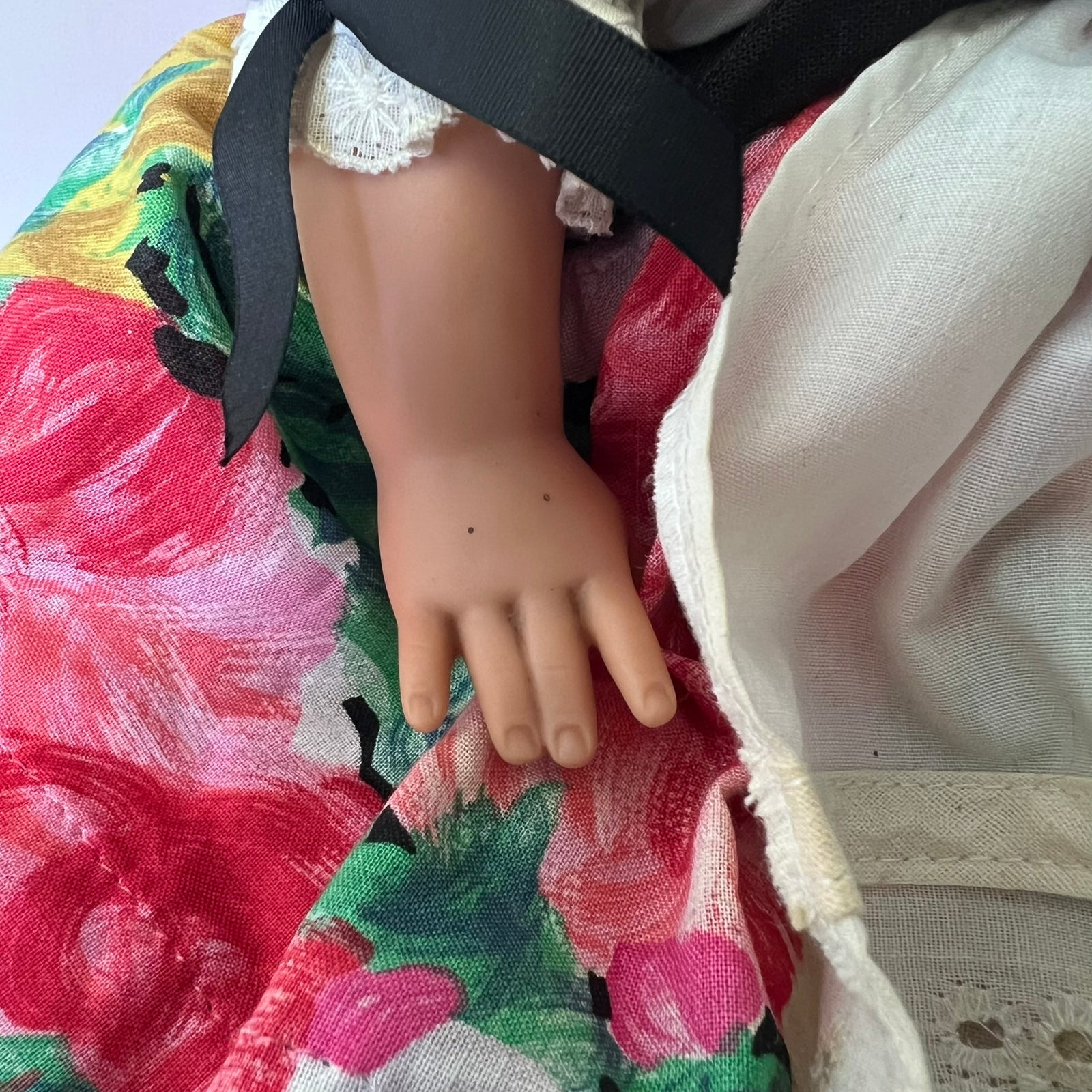 Close-up of a doll's hand resting on a colorful floral fabric, with part of a cream-colored dress and a black ribbon visible.