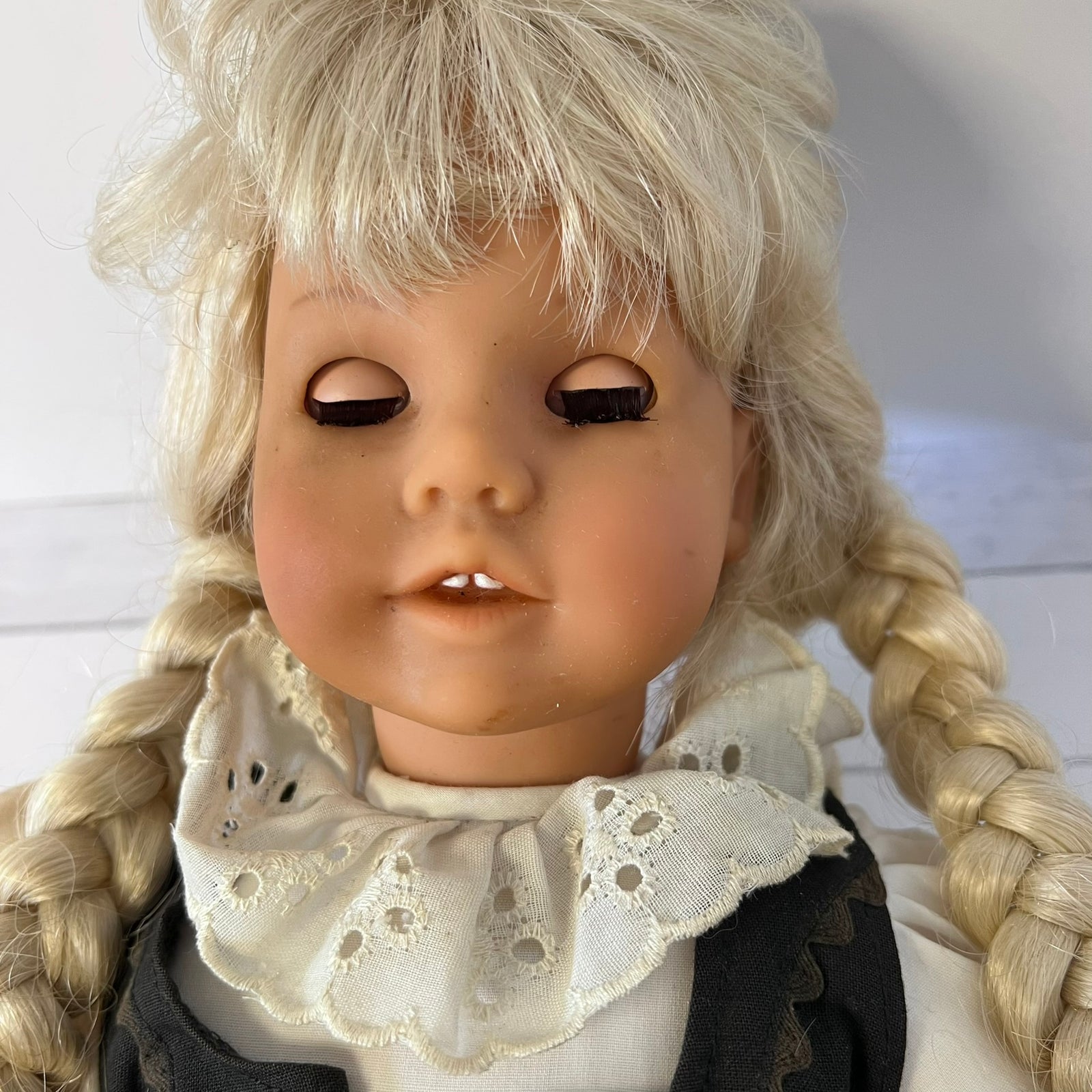 Vintage blonde doll with braided pigtails, a lace ruffled collar, and a dark dress, shown in a close-up shot.