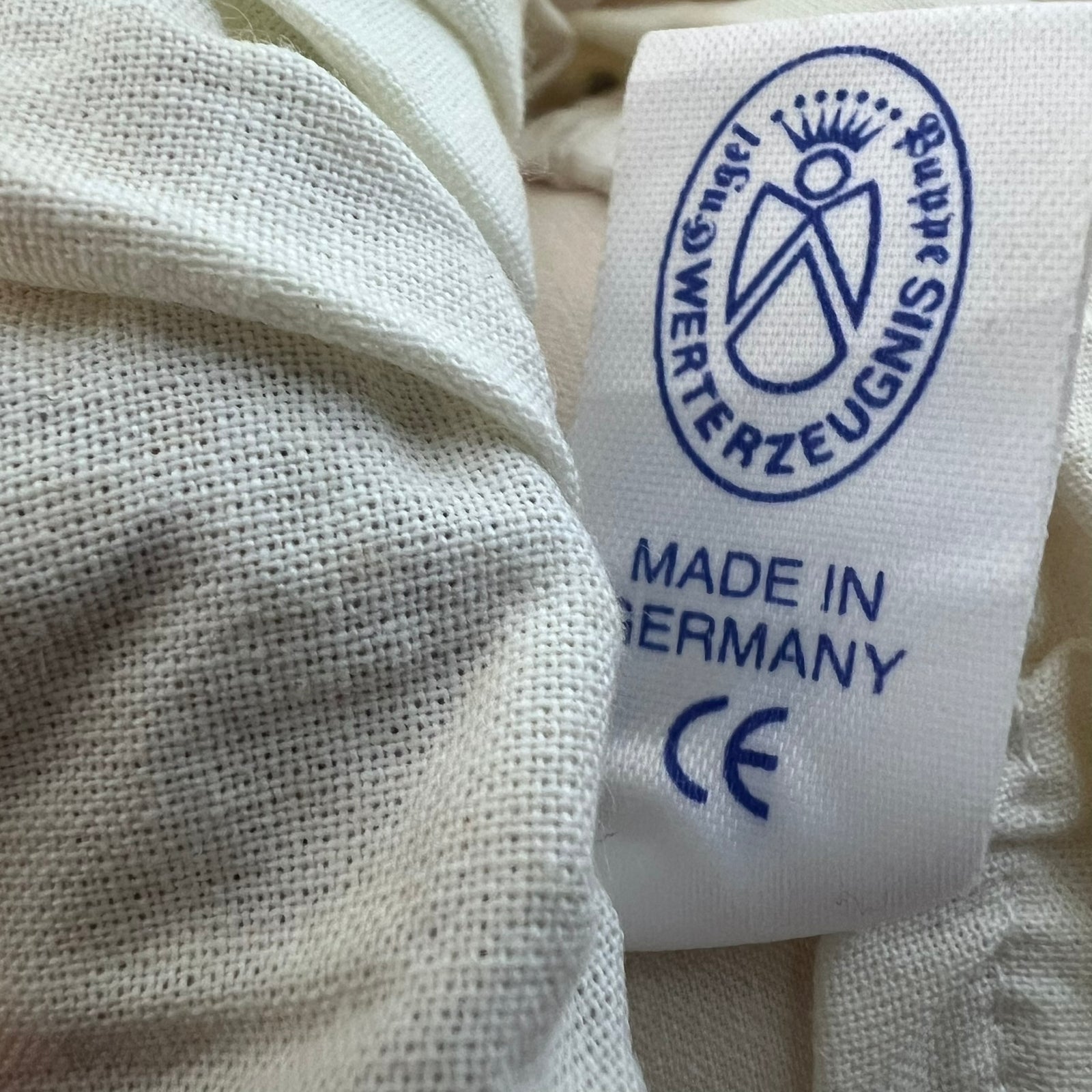 Close-up of a white woven fabric tag sewn onto off-white textured clothing, printed with a blue circular logo and the text 'MADE IN GERMANY' and 'CE'.